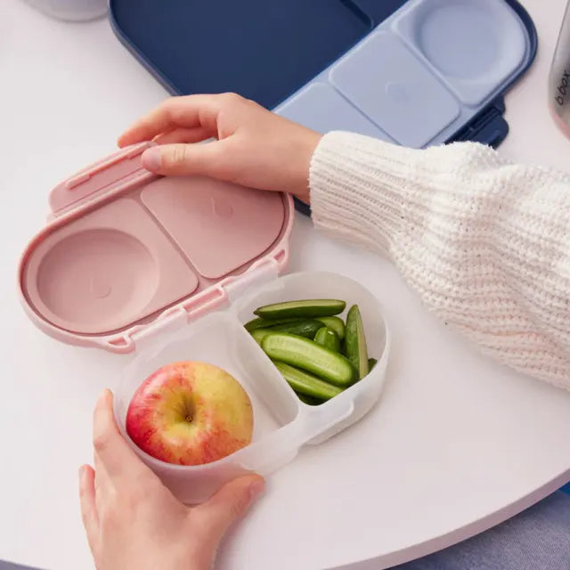 Pink and white compartmentalized snack container with an apple and green beans on a white surface.
