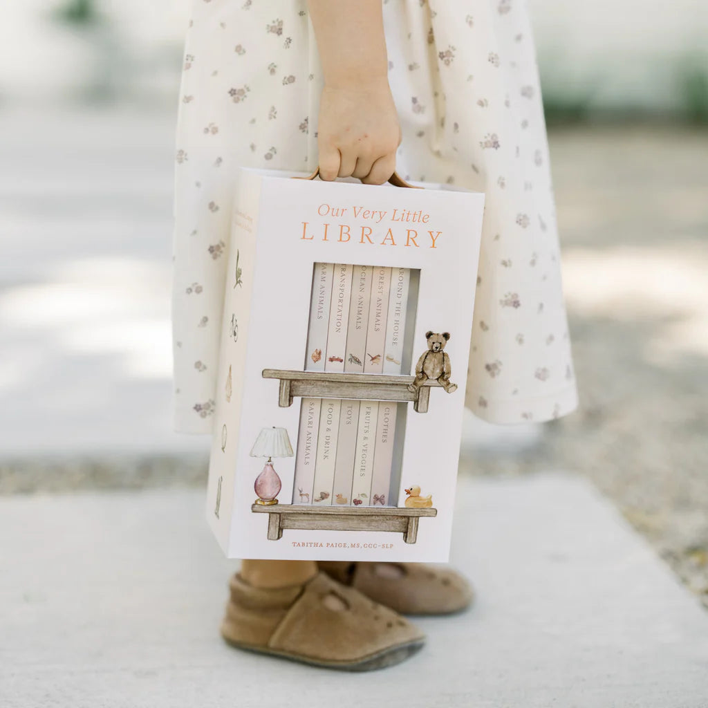 Person holding a book titled 'Our Very Little Library' with a blurred outdoor background