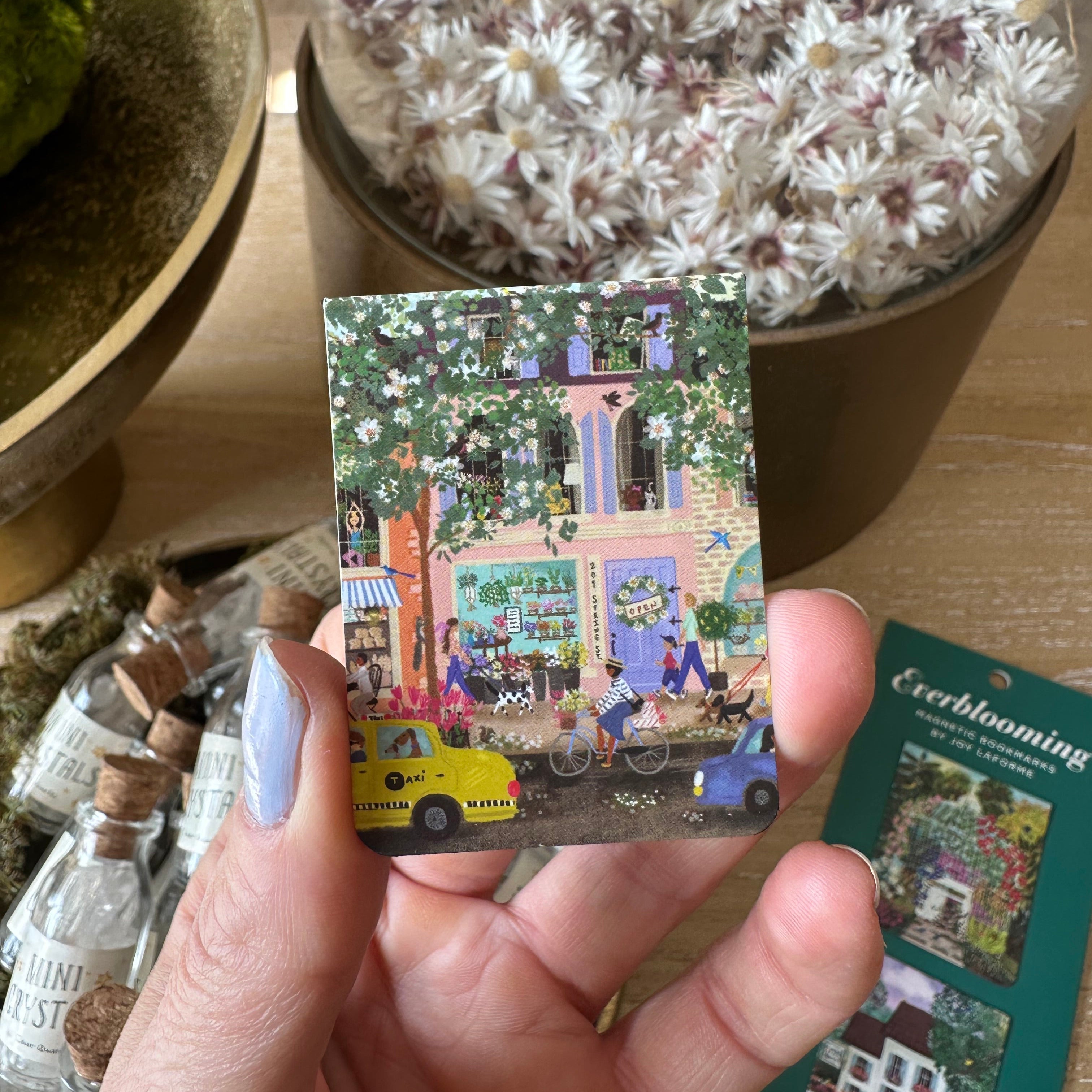 Hand holding a small card with a colorful design bookmark, surrounded by potted plants.