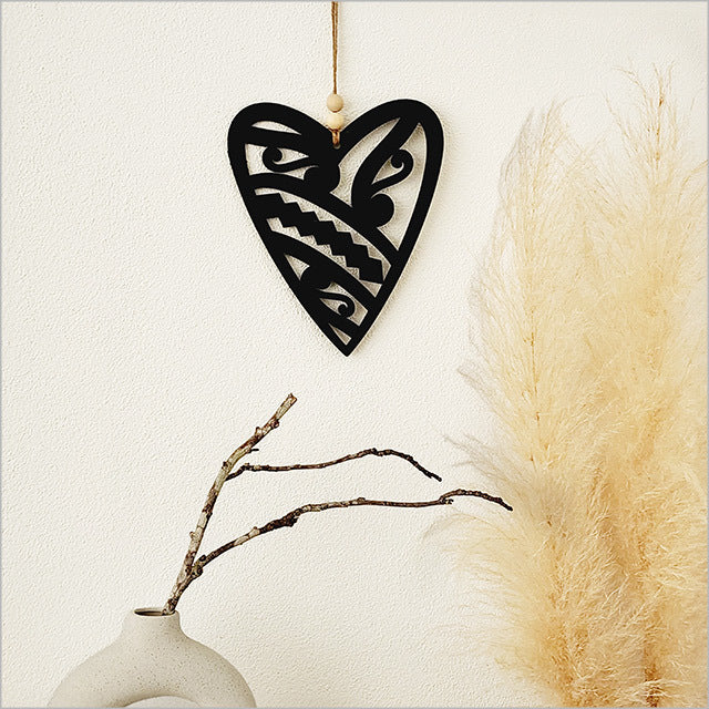 Black heart-shaped decorative item hanging against a neutral background with pampas grass and a vase.