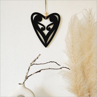 Black heart-shaped decorative item hanging on a white wall with pampas grass and a branch in the foreground.