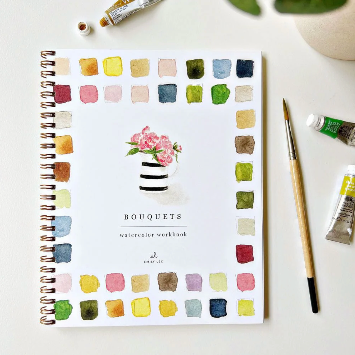 Watercolor workbook with color swatches and floral design on a white surface with art supplies.
