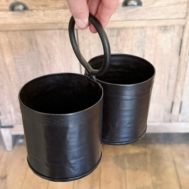 A hand holds a double metal canister with a single round handle, shown against a rustic wooden cabinet and floor background.
