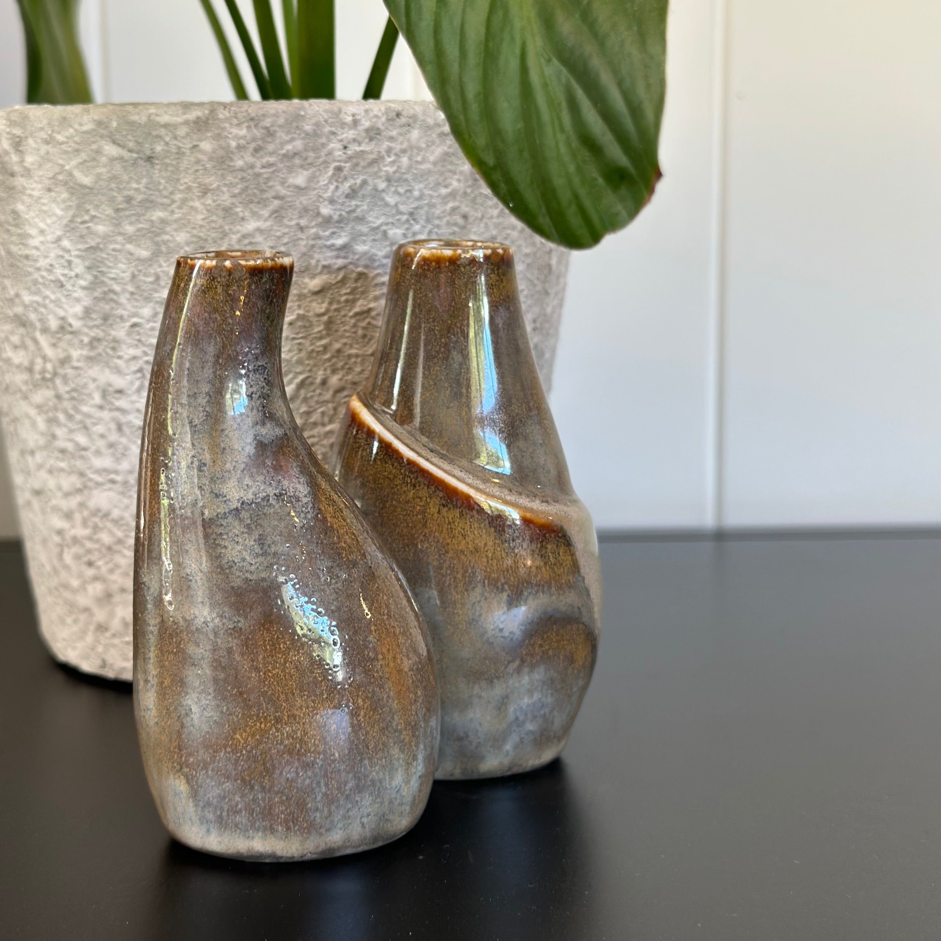 Two ceramic bottles with a textured finish on a dark surface, with a plant in the background.