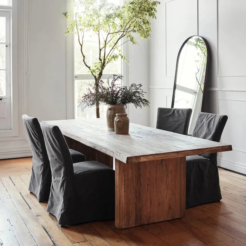 A rustic wooden dining table with a light weathered top and thick block legs, surrounded by six dark gray slipcovered chairs in a bright, modern dining room with hardwood floors and a tall floor mirror.