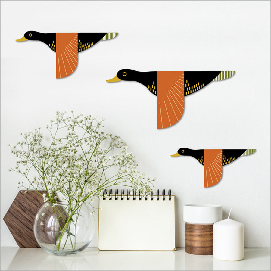 Decorative wall stickers of ducks on a white wall with a vase, notebook, and candle in the foreground.