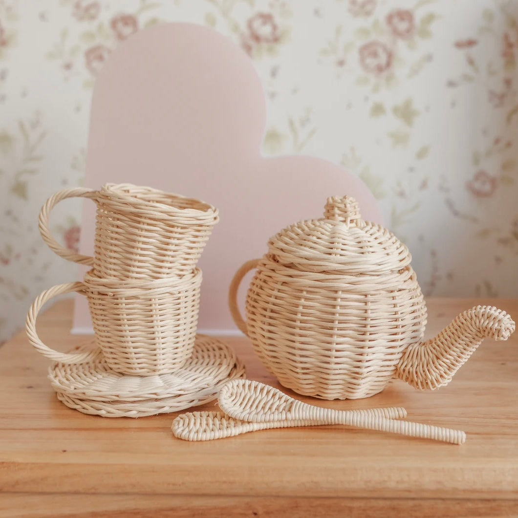 Kids rattan teapot, cup, and spoon set on a wooden surface with a floral-patterned background.