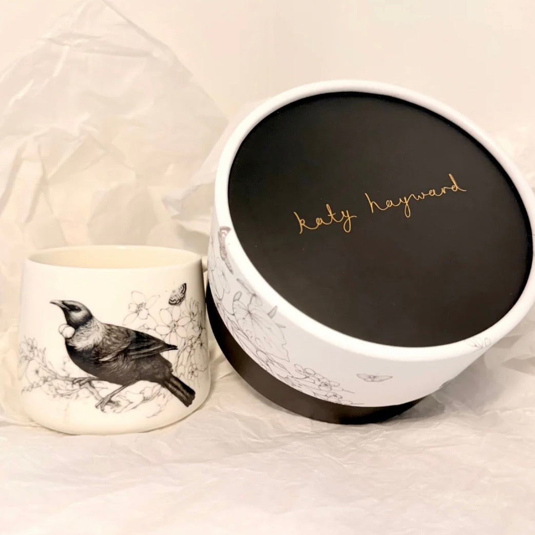 ceramic cup with a tui illustration by 'katy haywood', on a light background.