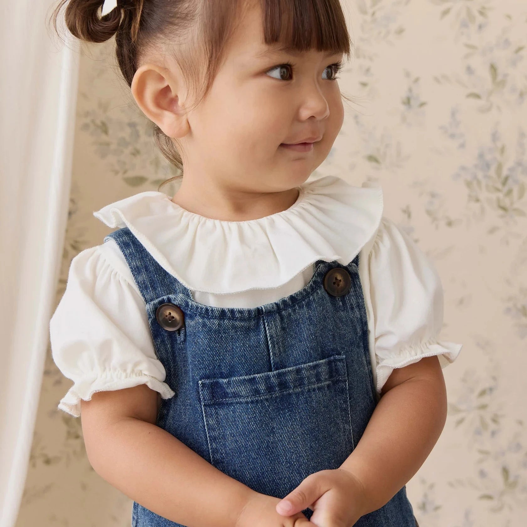 Child wearing a denim dress with a white blouse against a floral wallpaper background