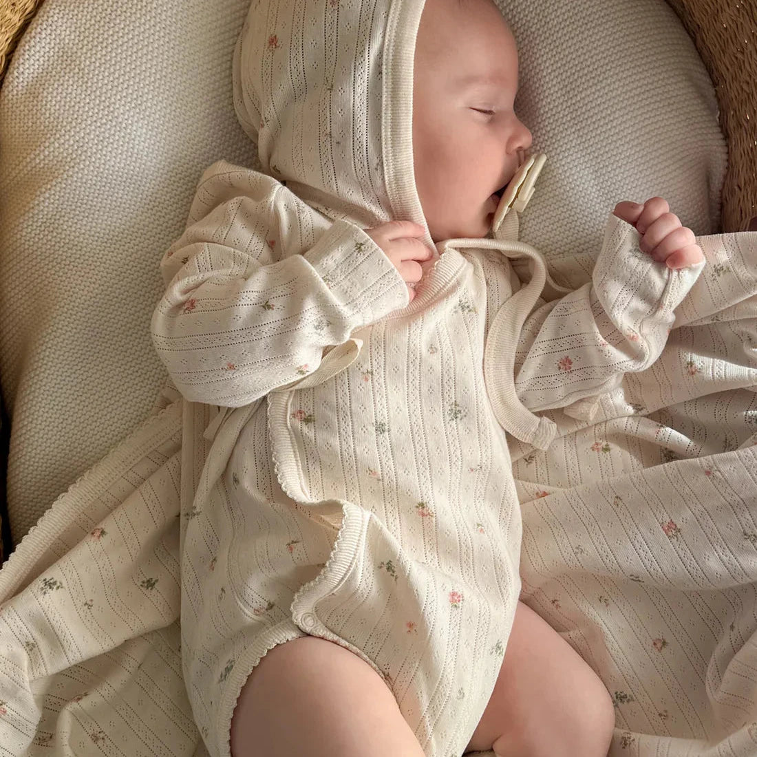Newborn baby in a white outfit with a hood, lying in a woven basket.