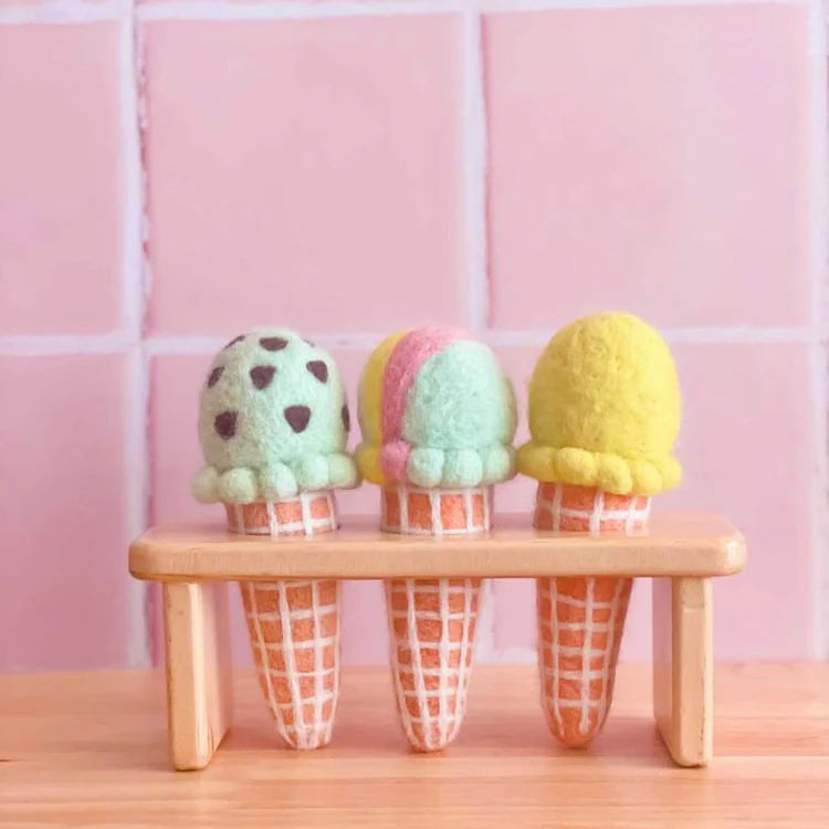 Three plush felt ice cream cones on a small wooden stool against a pink tiled wall.