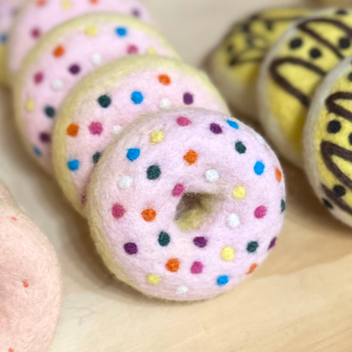 A close-up of several felt donut toys arranged on a light wood surface. The front donut is pale pink with multicolored sprinkle dots, and other donuts in the background include yellow with dark drizzle and pink with small red specks.