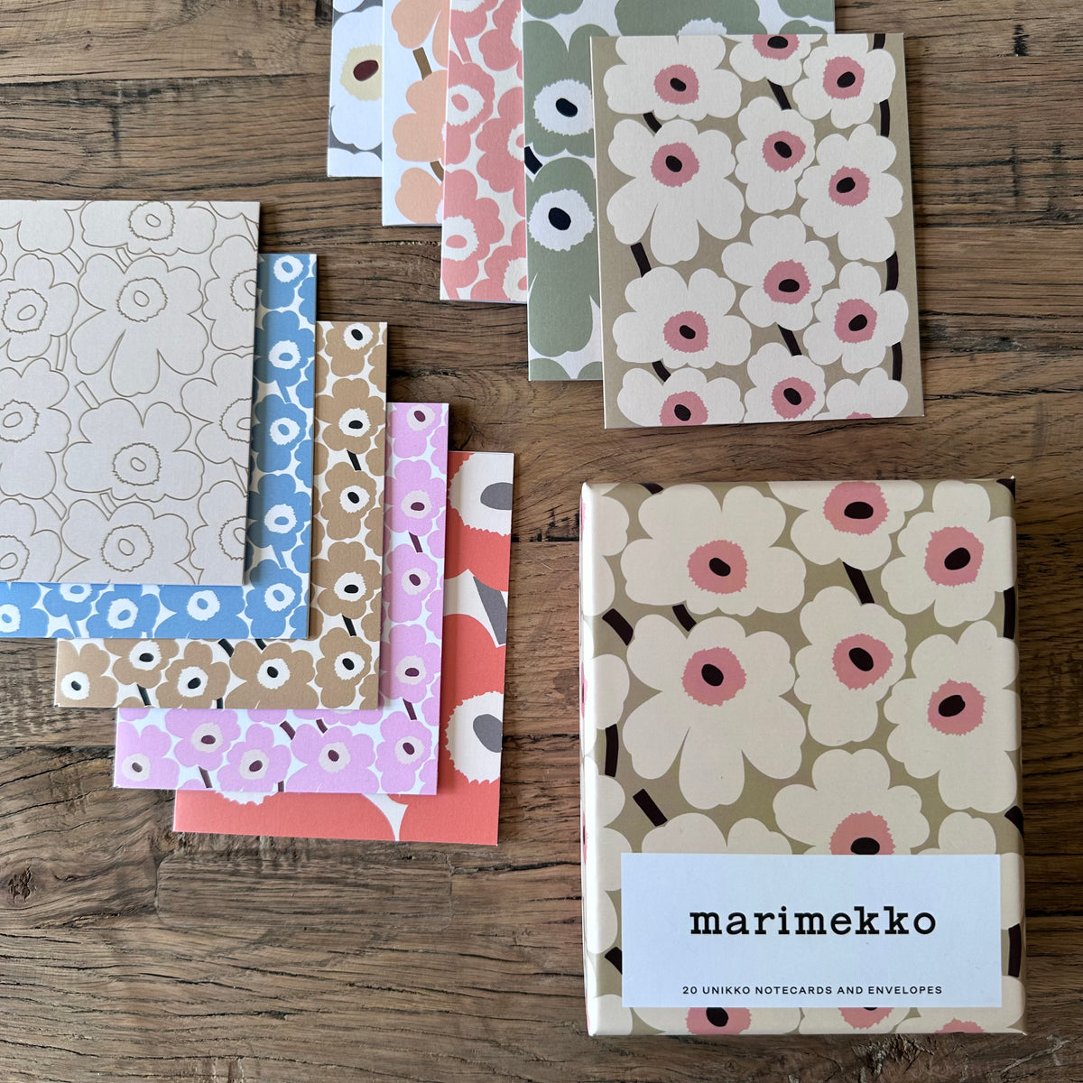 A set of Marimekko noteguards and envelopes arranged on a rustic wooden table. The set features multiple colorful designs with the iconic floral pattern in shades of pink, blue, brown, green, and orange on cream, beige, and pink backgrounds.