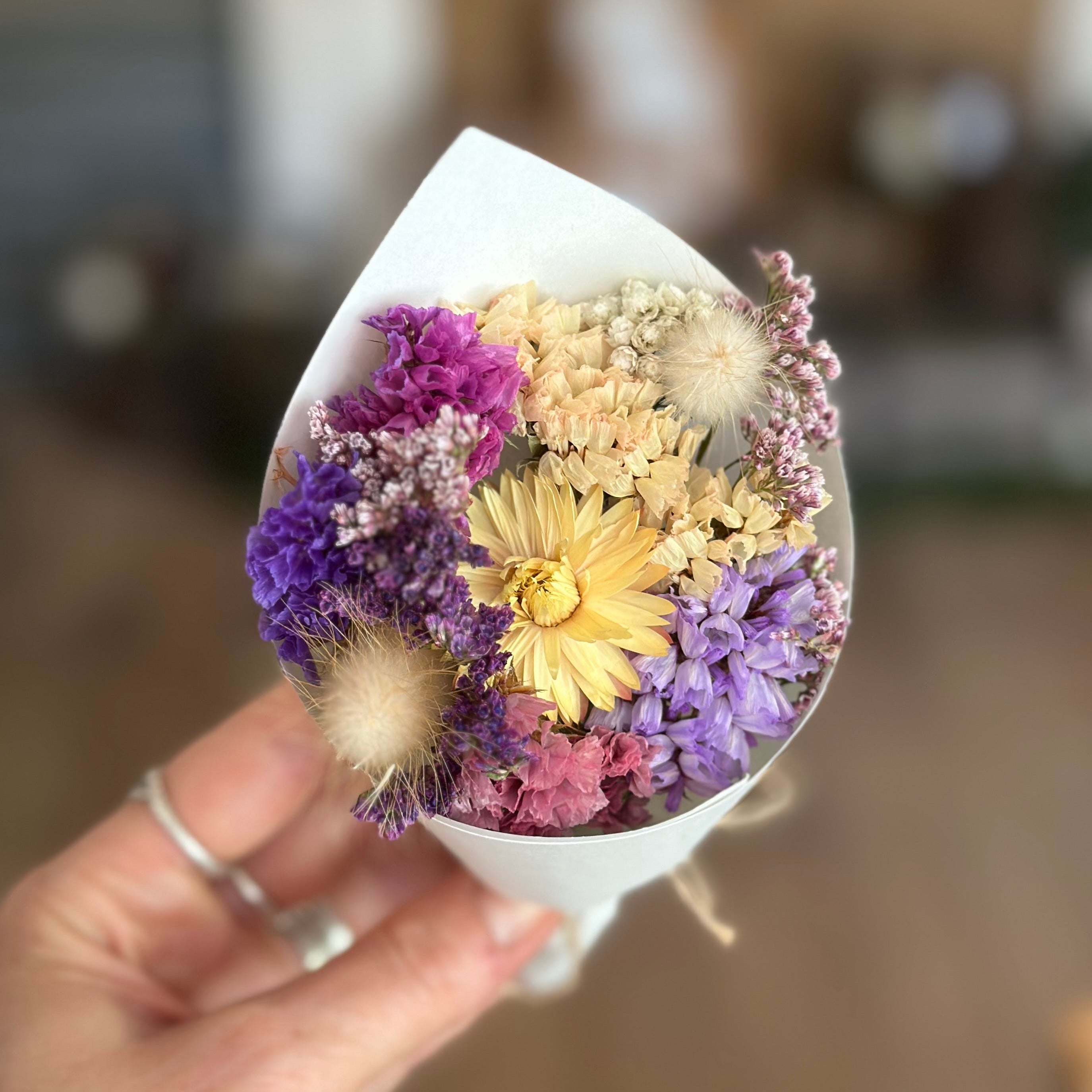 Hand holding a small bouquet of dried flowers in a white cone-shaped container against a blurred indoor background.
