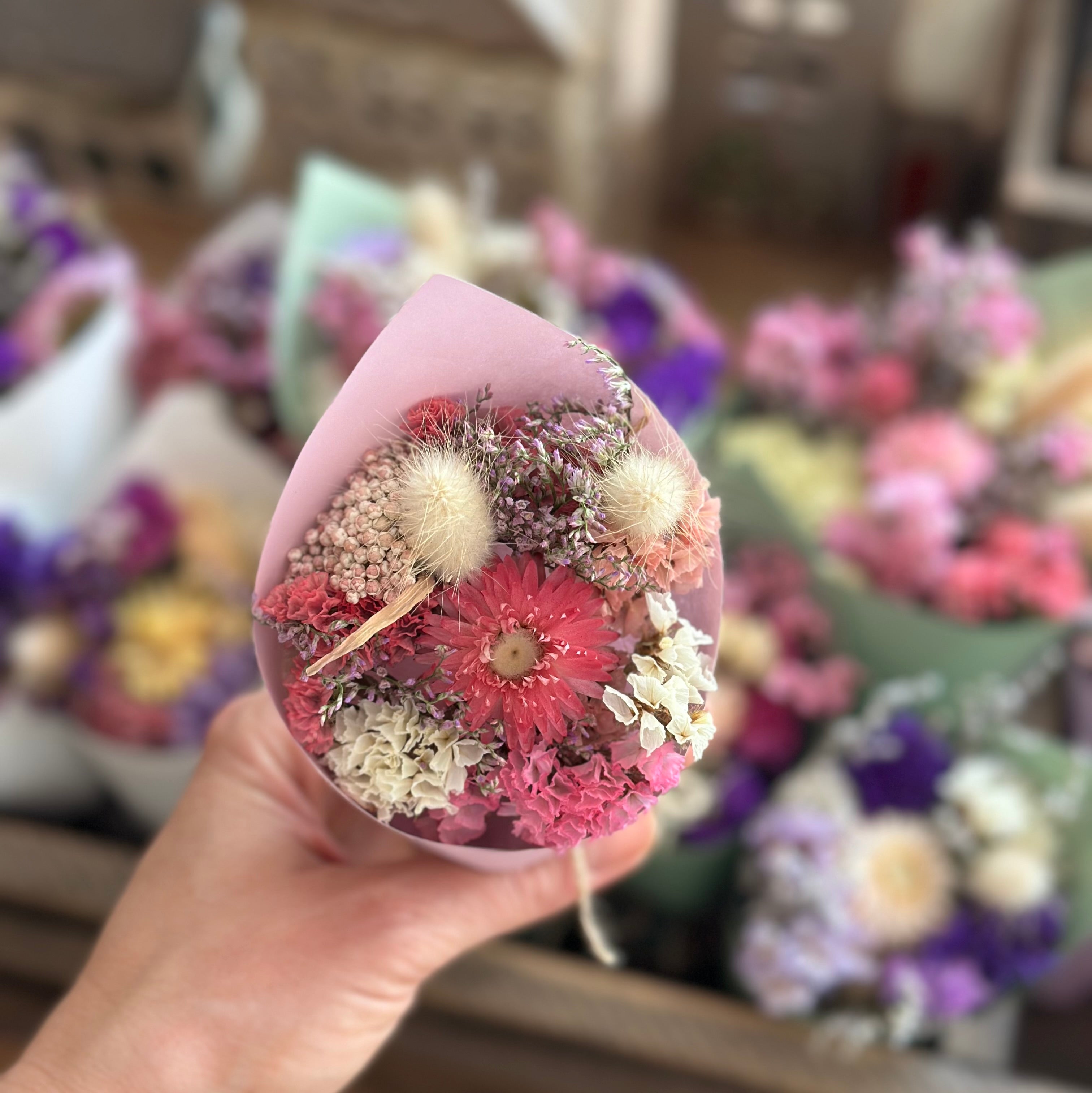 Hand holding a transparent container with dried flowers against a blurred background of more flower arrangements.