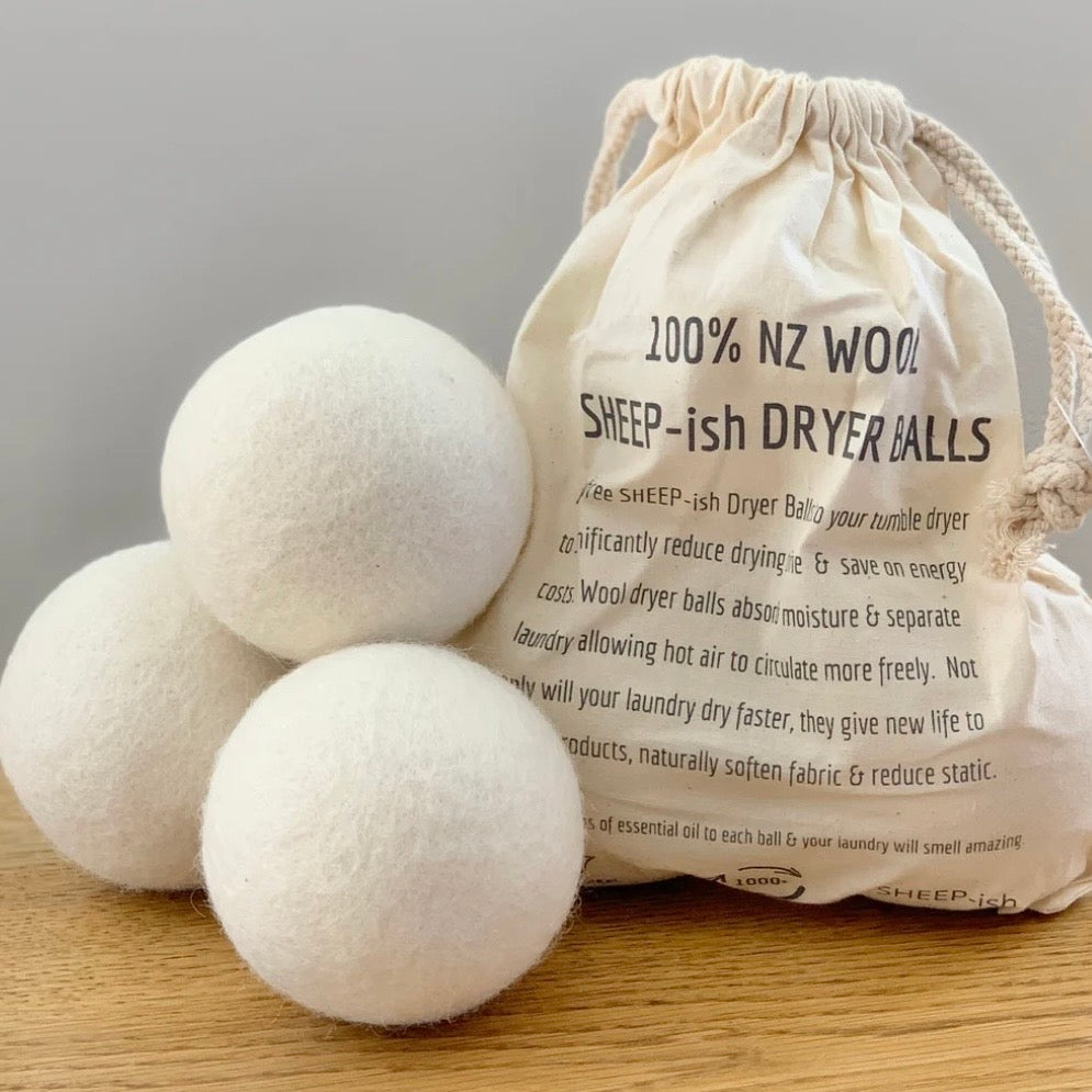 Three white dryer balls with a drawstring bag labeled '100% NZ Wool SHEEP-ish Dryer Balls' on a wooden surface.