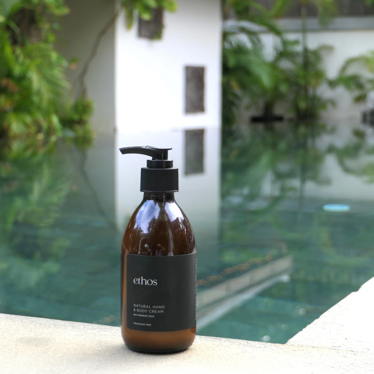 Brown bottle with black pump labeled 'ethos' on a ledge with a pool and greenery in the background