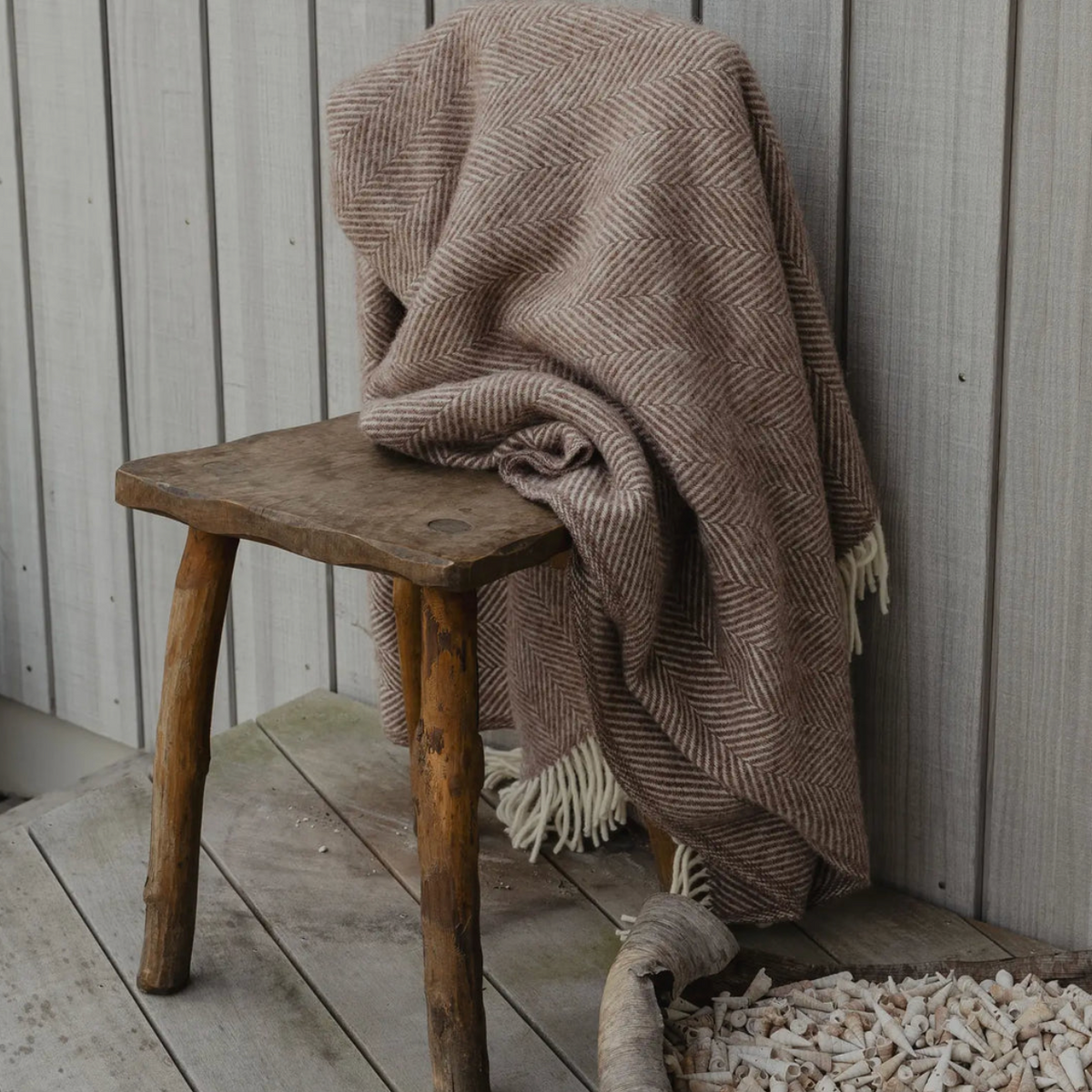 A beige and brown herringbone woven throw blanket with white fringe is draped over a rustic wooden stool on a weathered wooden deck against a gray wood-paneled wall.