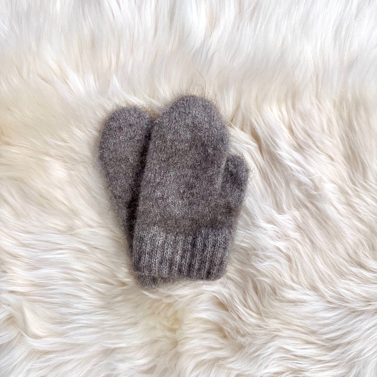 A pair of soft, merino and possom knit mittens in a heathered natural colour, laid flat on a fluffy white fur surface.