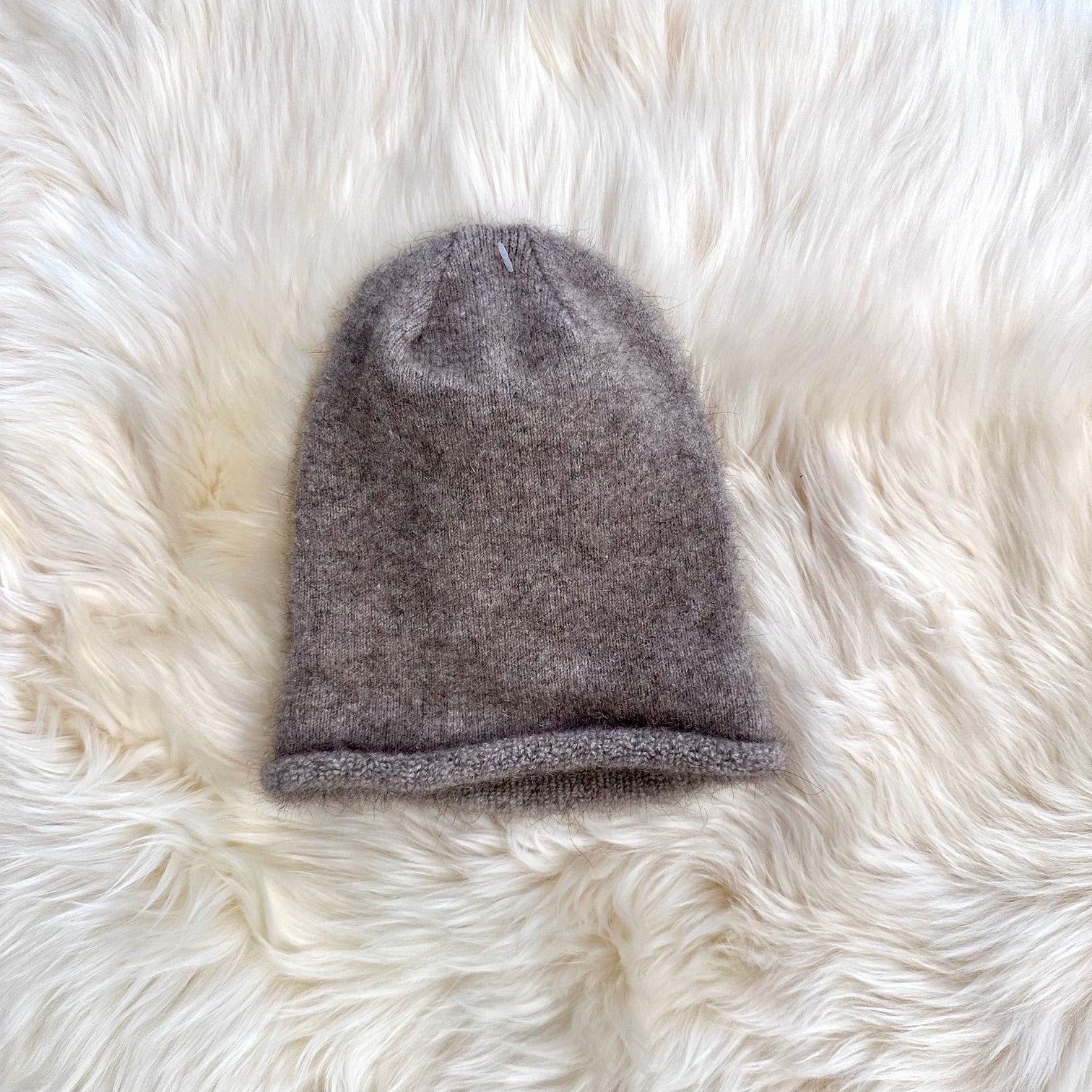 A single possum and merino knit beanie hat in a heathered taupe-gray color, laid flat on a white faux fur background.