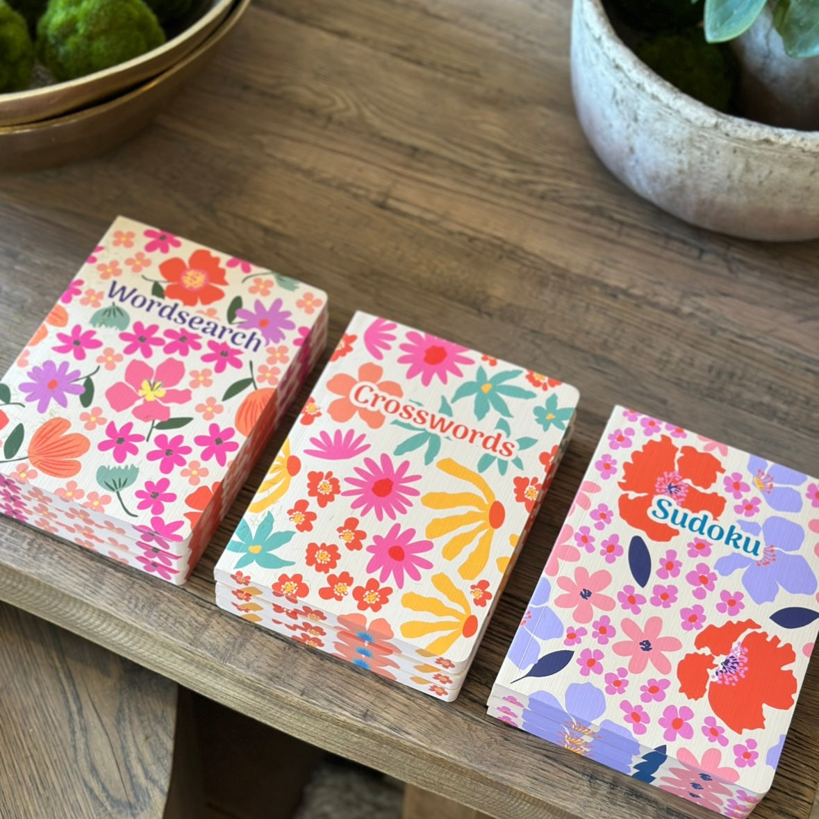 Three floral-themed puzzle books labeled 'Wordssearch', 'Crosswords', and 'Sudoku' on a wooden surface.