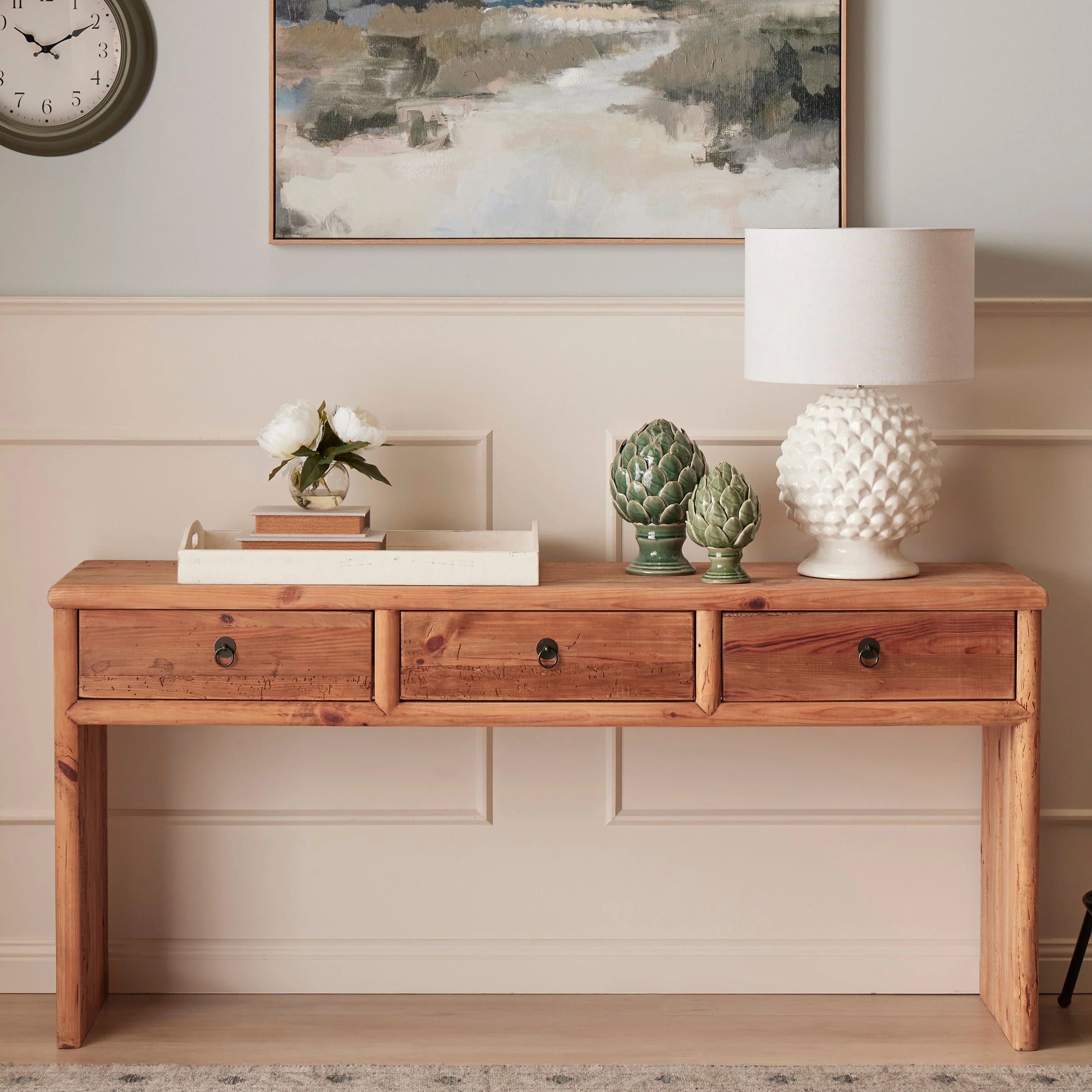 Wooden console table with decorative items against a wall with a clock and painting.