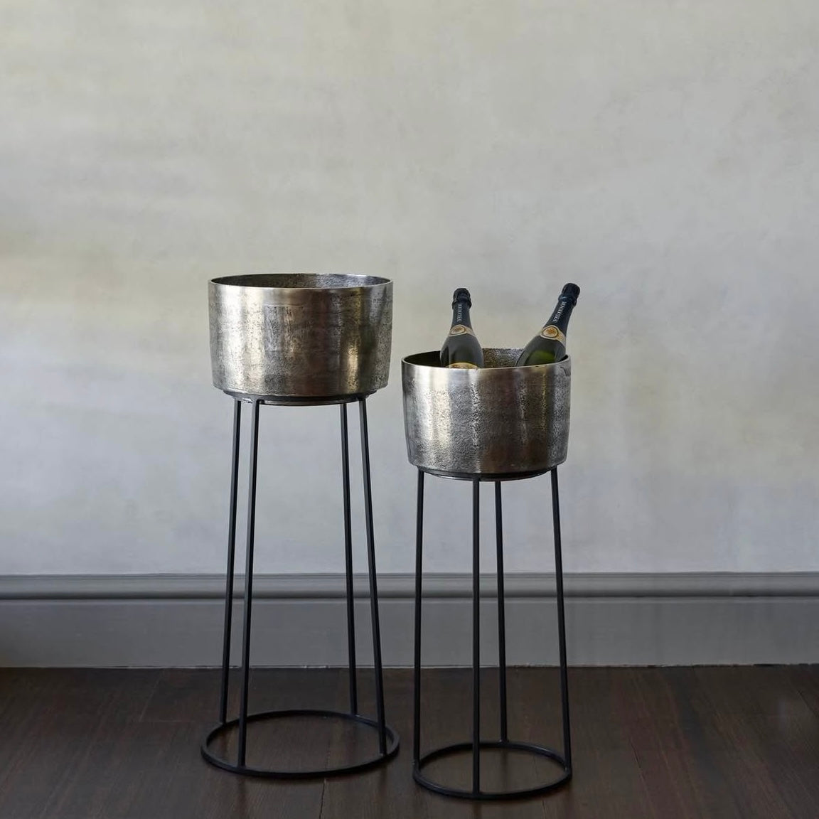 Two metallic planters with black legs on a wooden floor against a plain wall.