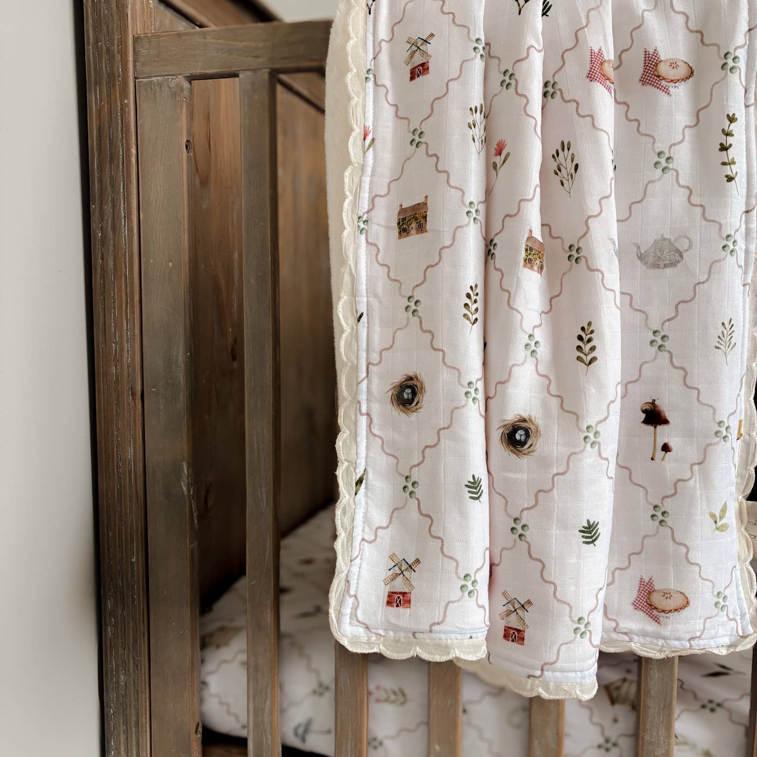 A close-up of a white baby blanket draped over a wooden crib, featuring a beige trellis pattern with embroidered woodland motifs like a fox, mushroom, and bird.