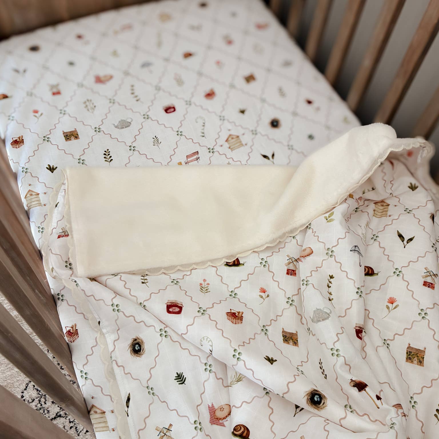 A close-up of a baby bedding set inside a wooden crib, featuring a quilted mattress cover and a matching folded blanket with a cream background and a whimsical multicolor print.