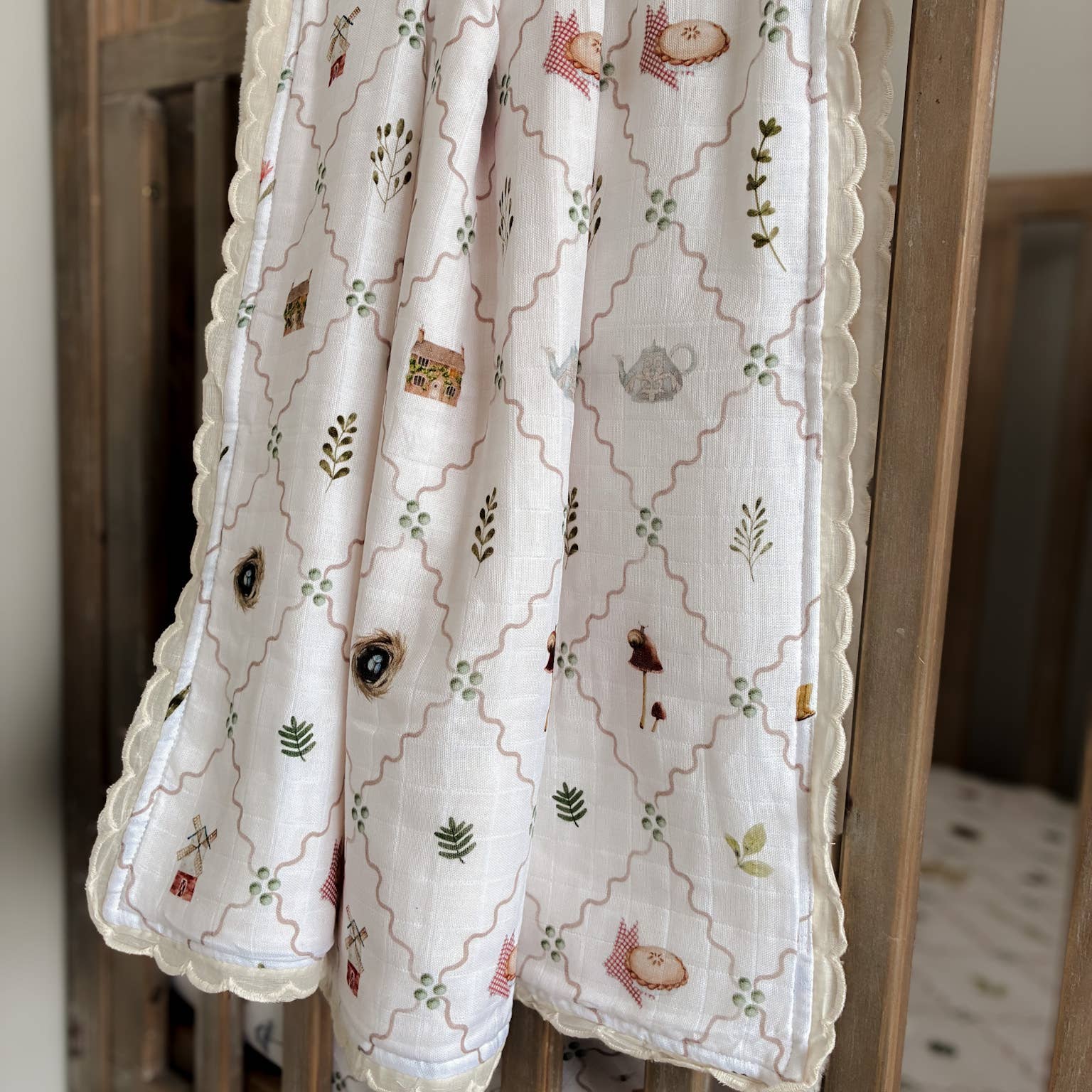 A white muslin baby blanket with a scalloped edge, featuring a printed woodland pattern of foxes, birds, leaves, and a trellis design, draped over the side of a wooden crib.