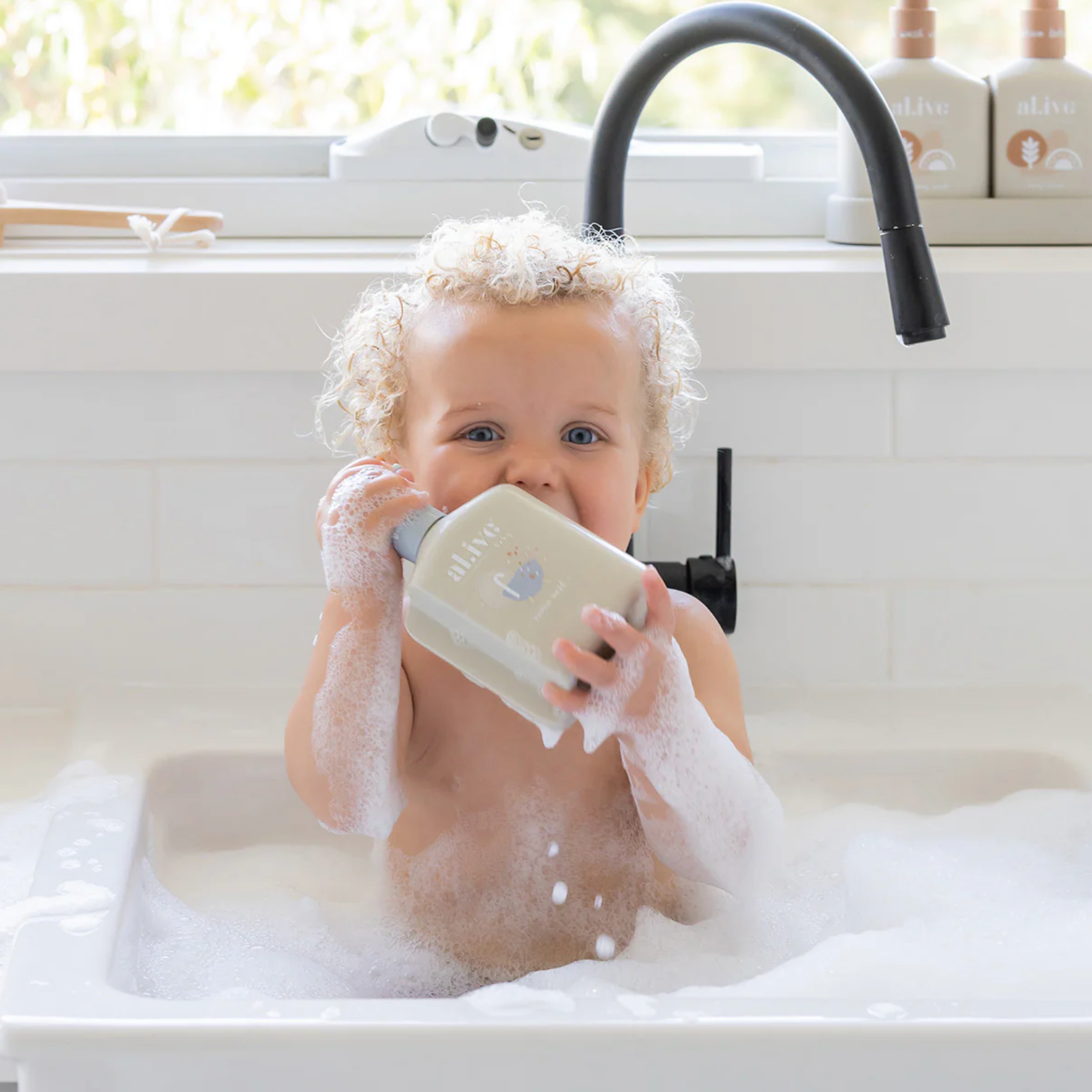 A baby with curly blonde hair is sitting in a white bathtub filled with soapy water, holding a beige bottle labeled 'olive' in front of their face. A black faucet is visible in the background above the tub.