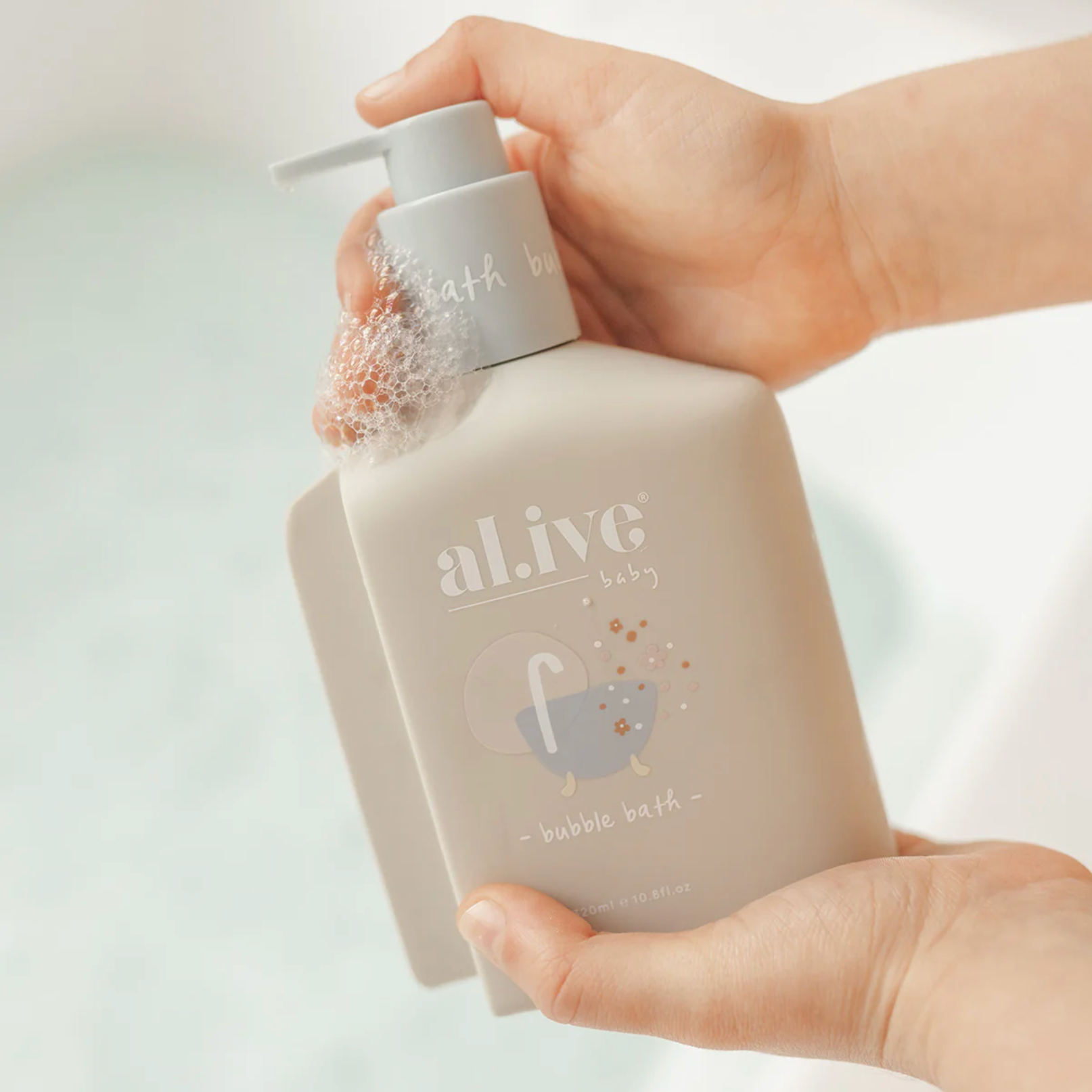 A person holds a frosted beige bottle of al ive baby bubble bath with a white pump dispenser, covered in soapy bubbles, against a soft, bright bathroom background.