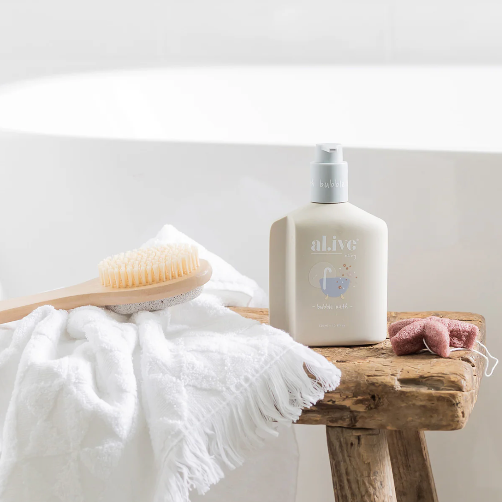 An 'alive' baby shower bottle with a pump dispenser, styled on a rustic wooden stool next to a white bath towel, a wooden baby brush, and a pink plush toy.