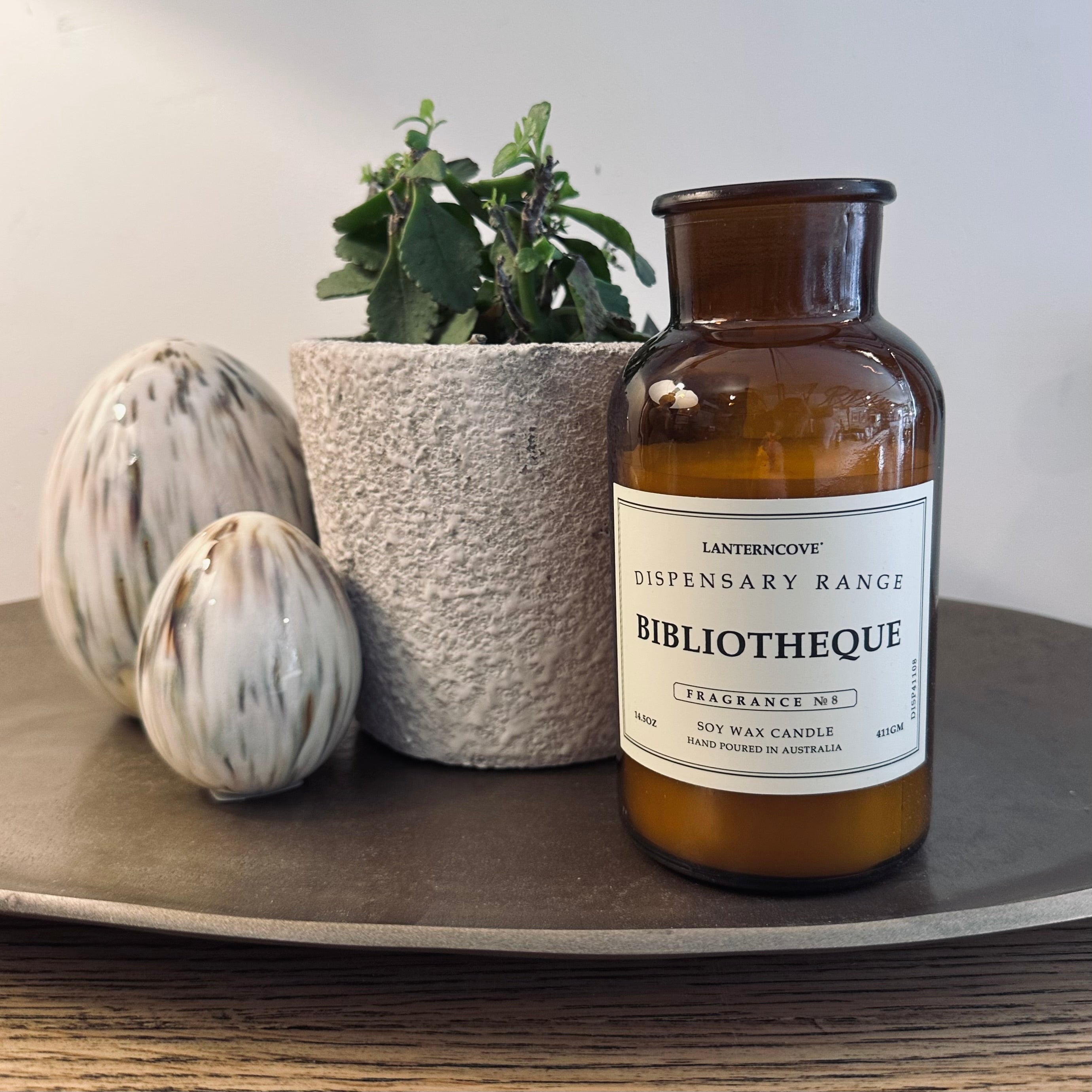 A soy wax candle in an amber glass bottle with a white label reading 'BIBLIOTHEQUE' from 'LANTERNBROKE DISPENSARY RANGE', styled on a dark tray with a potted plant and decorative ceramic objects against a neutral background.
