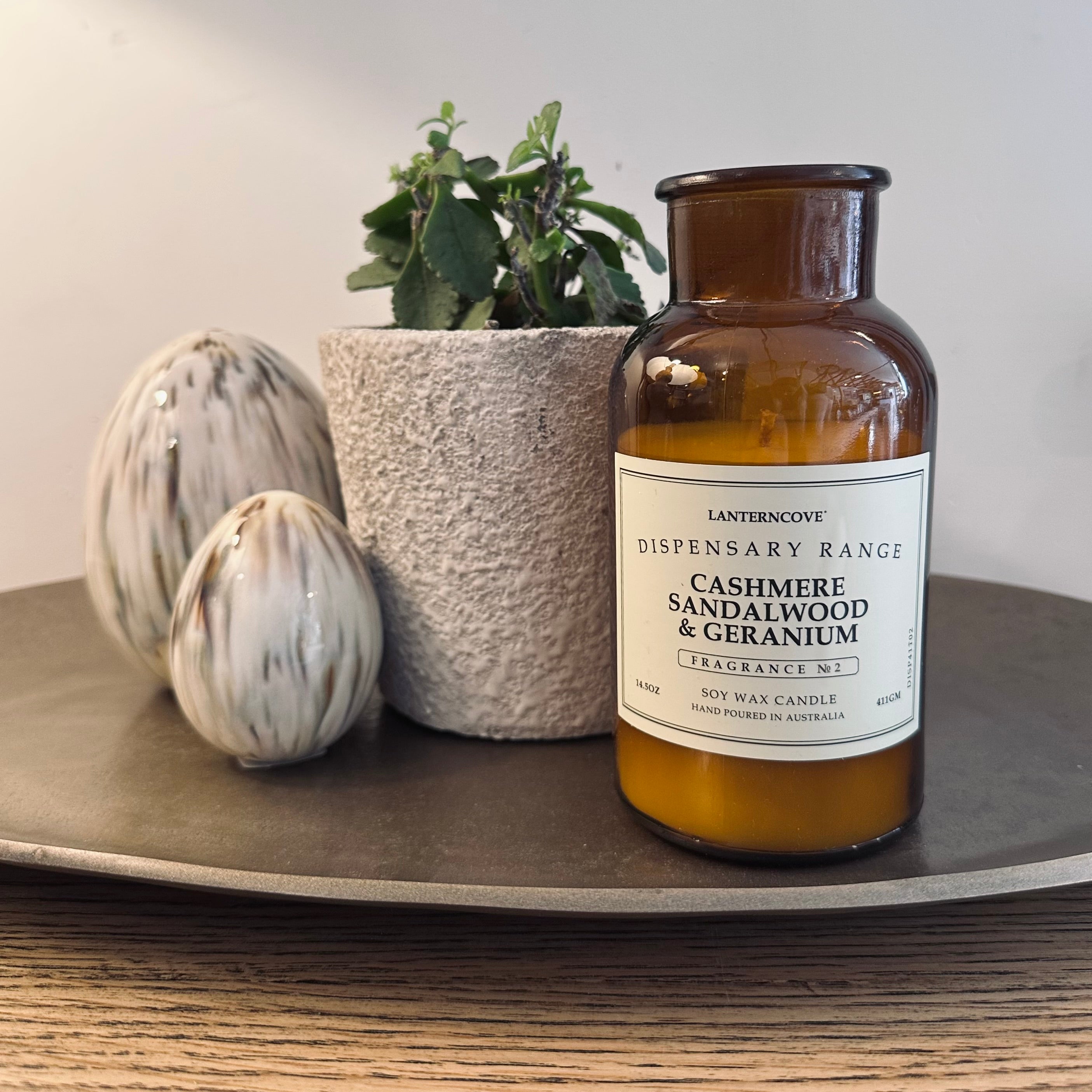 An amber glass soy wax candle jar with a white label reading 'LANTERNOCOE DISPENSARY RANGE CASHMERE SANDALWOOD & GERANIUM' sits on a round gray tray. The tray is styled with two white speckled decorative eggs and a small potted green plant against a neutral beige wall.