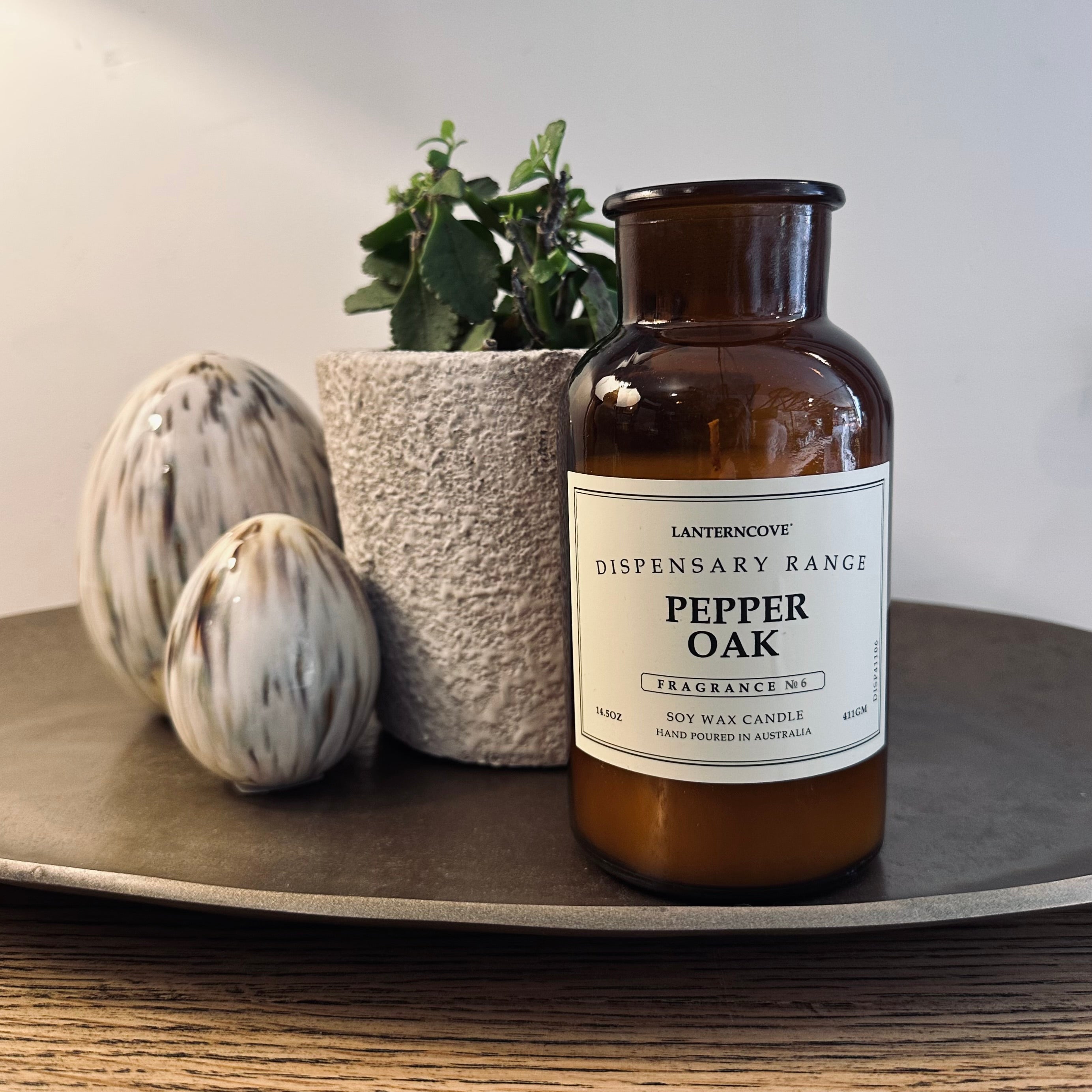 An amber glass candle jar labeled 'Lantern Cove Dispensary Range Pepper Oak Fragrance No. 5' sits on a round tray beside a small potted plant and two decorative eggs against a plain light background.