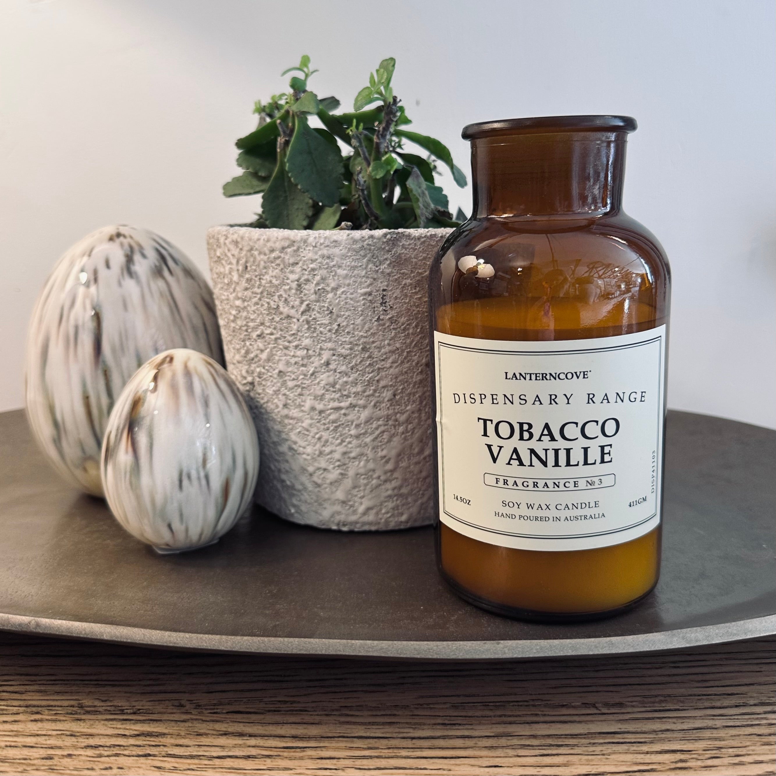 An amber glass scented candle bottle labeled 'Lanterncove Dispensary Range Tobacco Vanille Fragrance No. 1' sits on a round tray beside a potted green plant and two decorative ceramic eggs against a neutral wall.