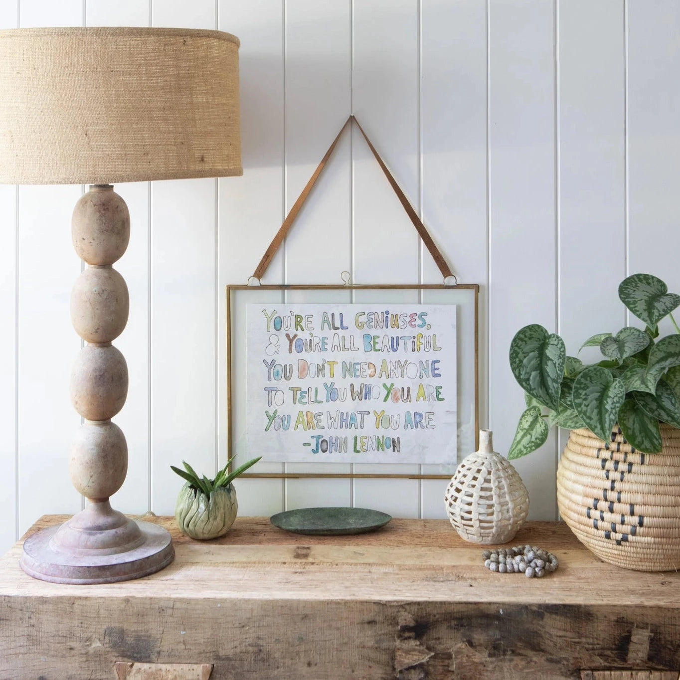 A framed John Lennon quote print hangs on a white paneled wall above a rustic wooden shelf styled with a tall woven lamp base, a potted green plant, and other home decor accessories.