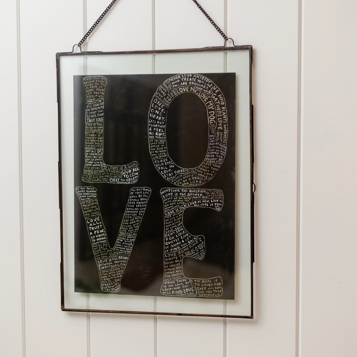 Framed wall art hanging on a white paneled wall, featuring the word LOVE in large letters filled with small text on a dark background.