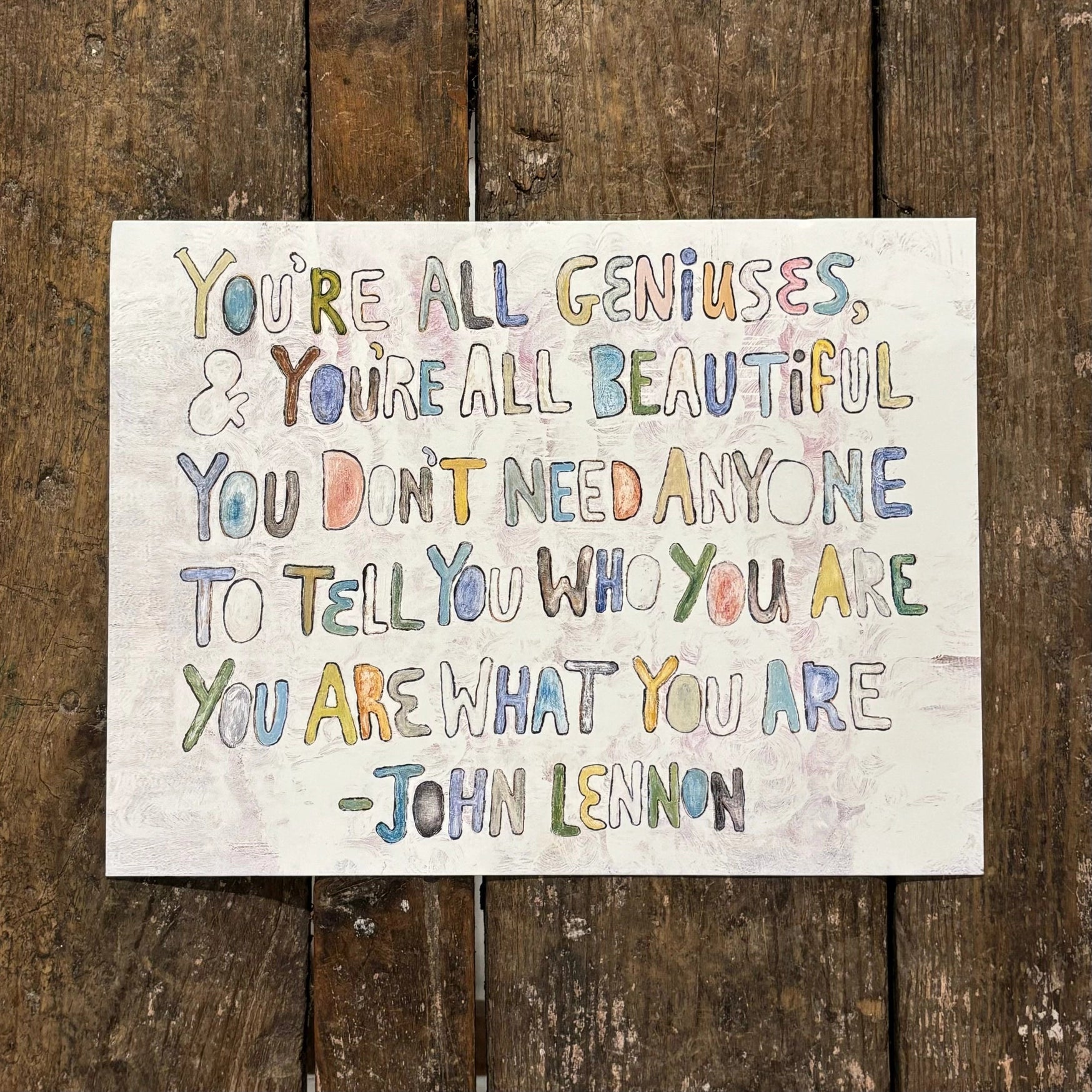 A square white art print featuring a John Lennon quote in colorful, hand-drawn lettering, displayed flat on a rustic dark brown wooden plank background.