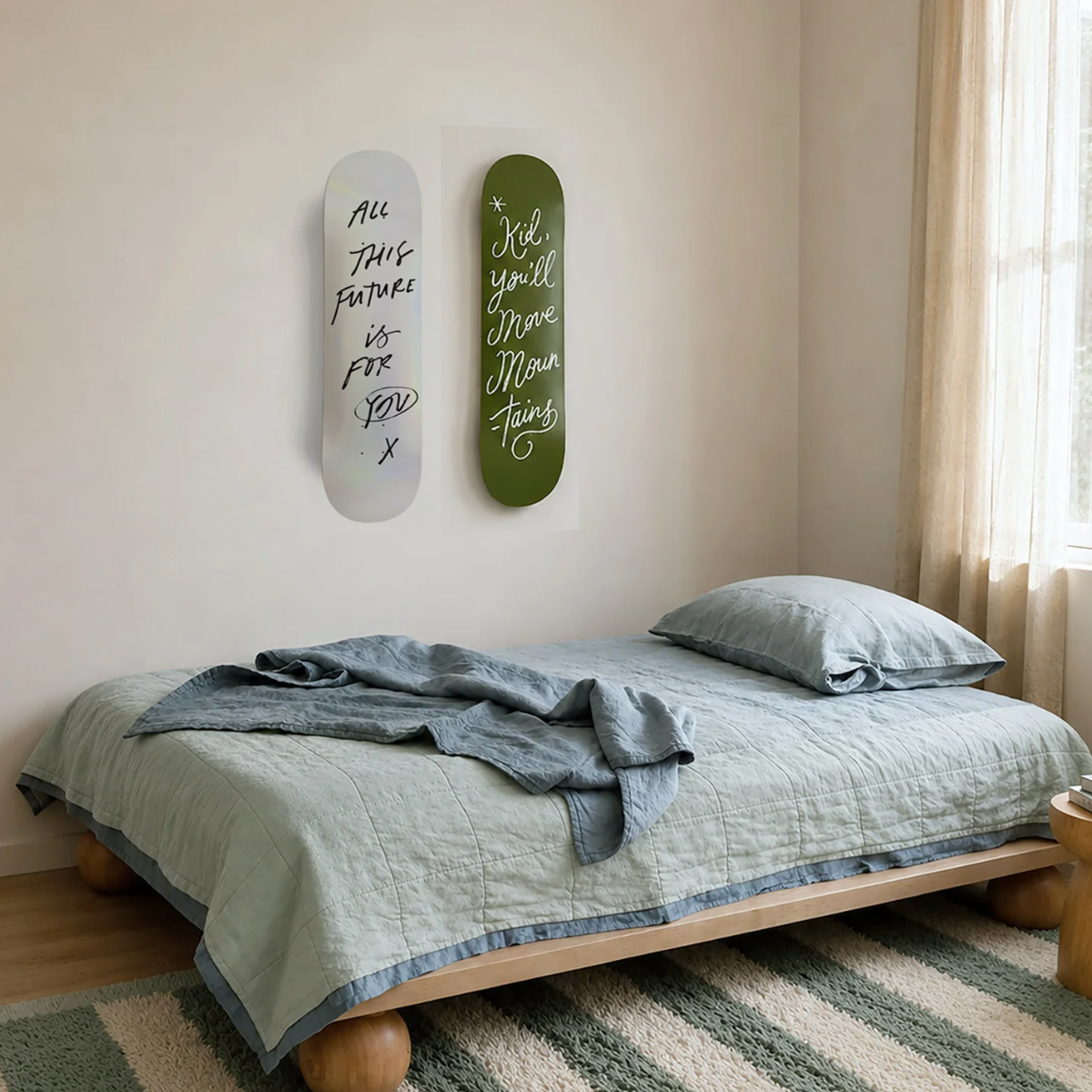 A minimalist bedroom scene with a low wooden bed frame dressed in pale sage bedding and a gray throw blanket. Two skateboard-style wall art pieces with handwritten quotes hang above the bed on a cream wall, with a window and sheer curtains to the right.