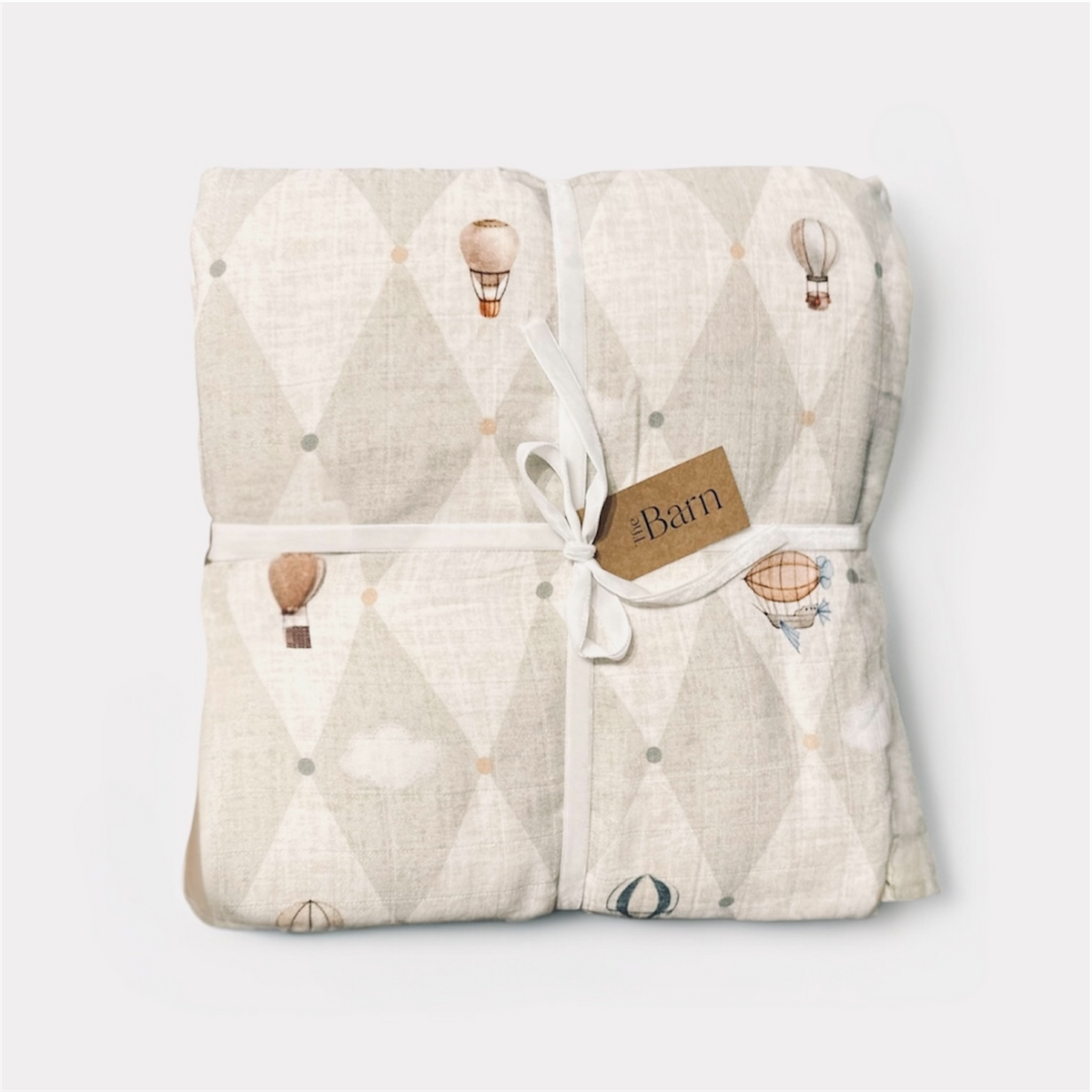 A folded baby blanket with a light beige and off-white diamond pattern, decorated with small hot air balloon and cloud illustrations. The blanket is tied with a white ribbon and a brown paper tag that reads '4Barn'.