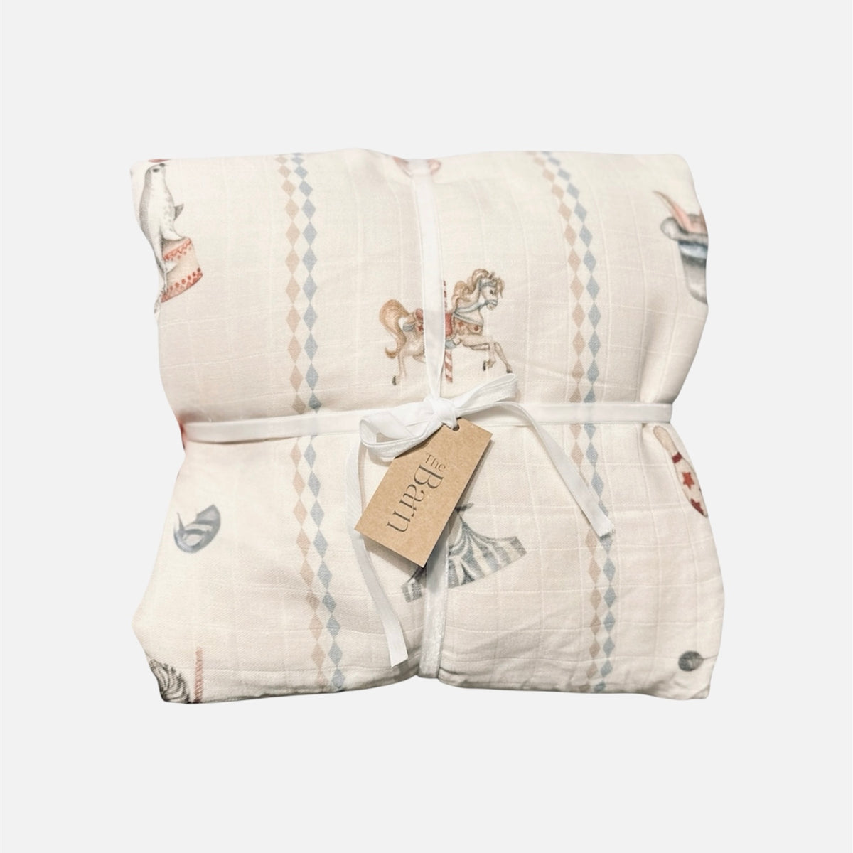 A folded muslin blanket with a vintage carousel print, tied with a white ribbon and a paper brand tag that reads 'the barn'. The blanket has a warm cream base with muted blue, pink, and brown illustrations of carousel horses and vertical diamond-patterned stripes.