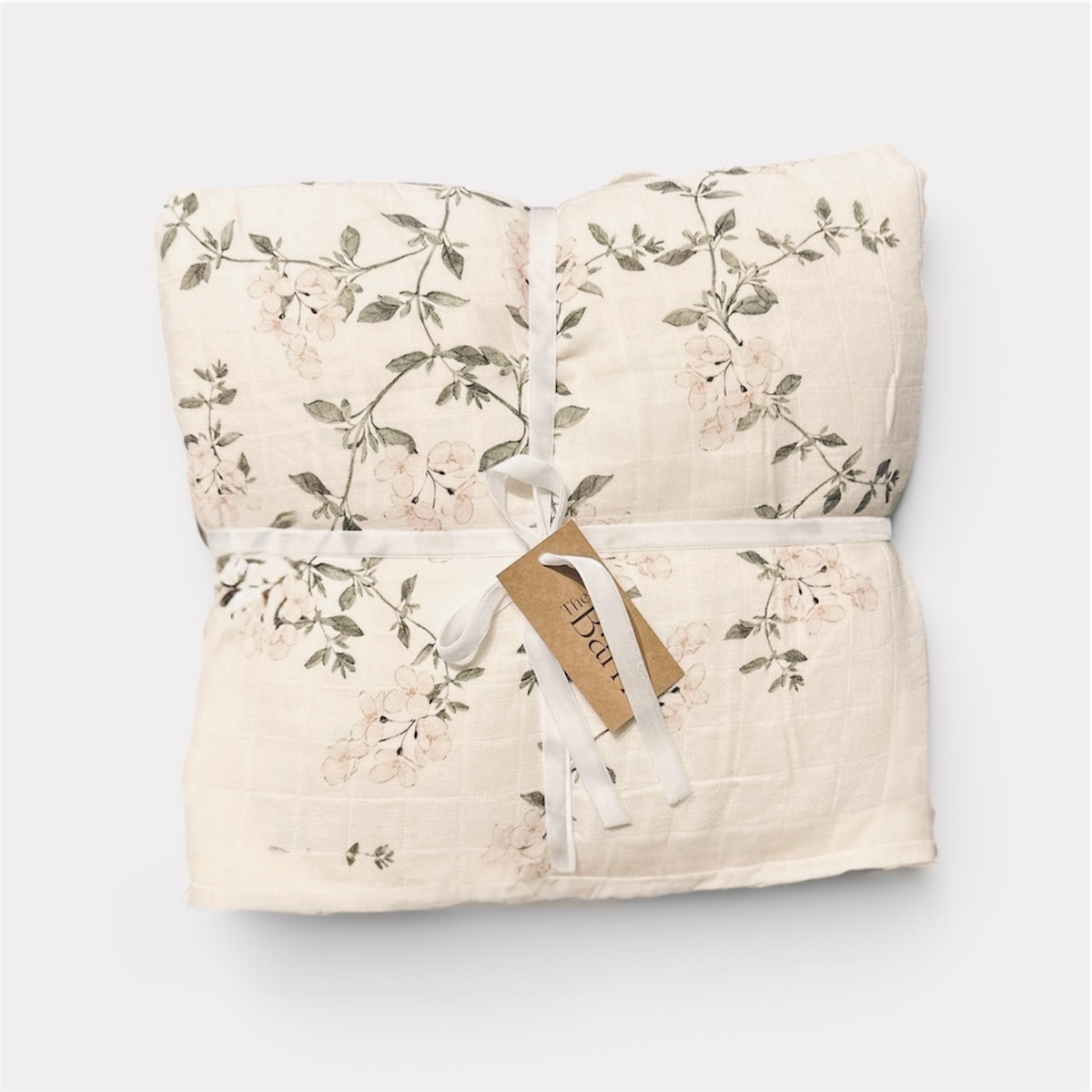 A folded quilted cotton blanket with a delicate floral print in soft pink and muted green on an off-white background, tied with a white ribbon and a kraft paper tag.