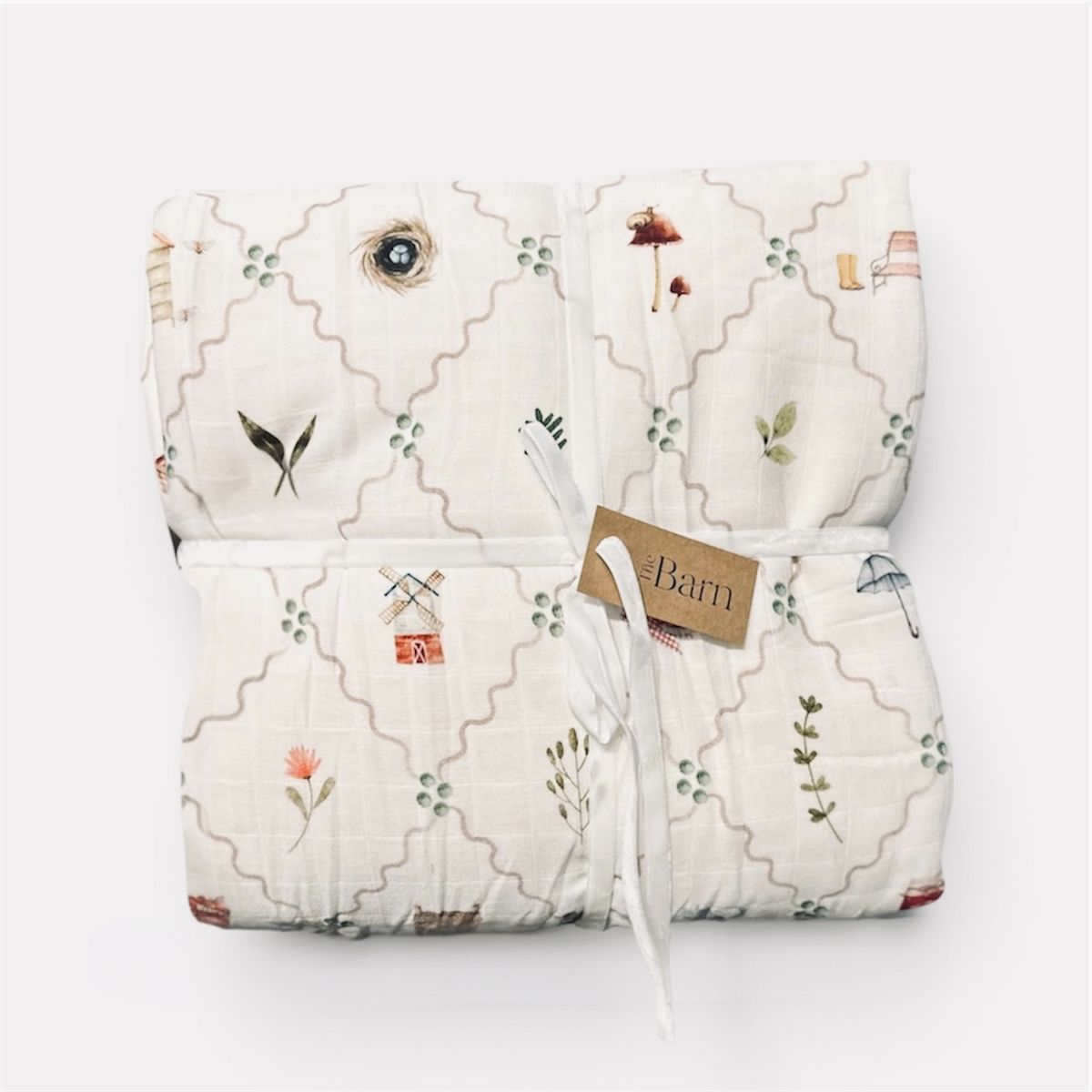 A folded muslin blanket with a beige trellis pattern and small whimsical illustrations, tied with a white ribbon and a paper tag that reads 'Barn'.