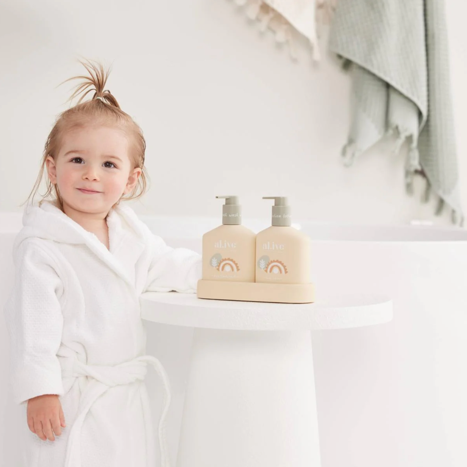 A young child in a white bathrobe stands beside a small white table holding two beige pump bottles with a rainbow graphic. The scene is softly lit in a bright, minimal bathroom-like setting with a towel hanging in the background.