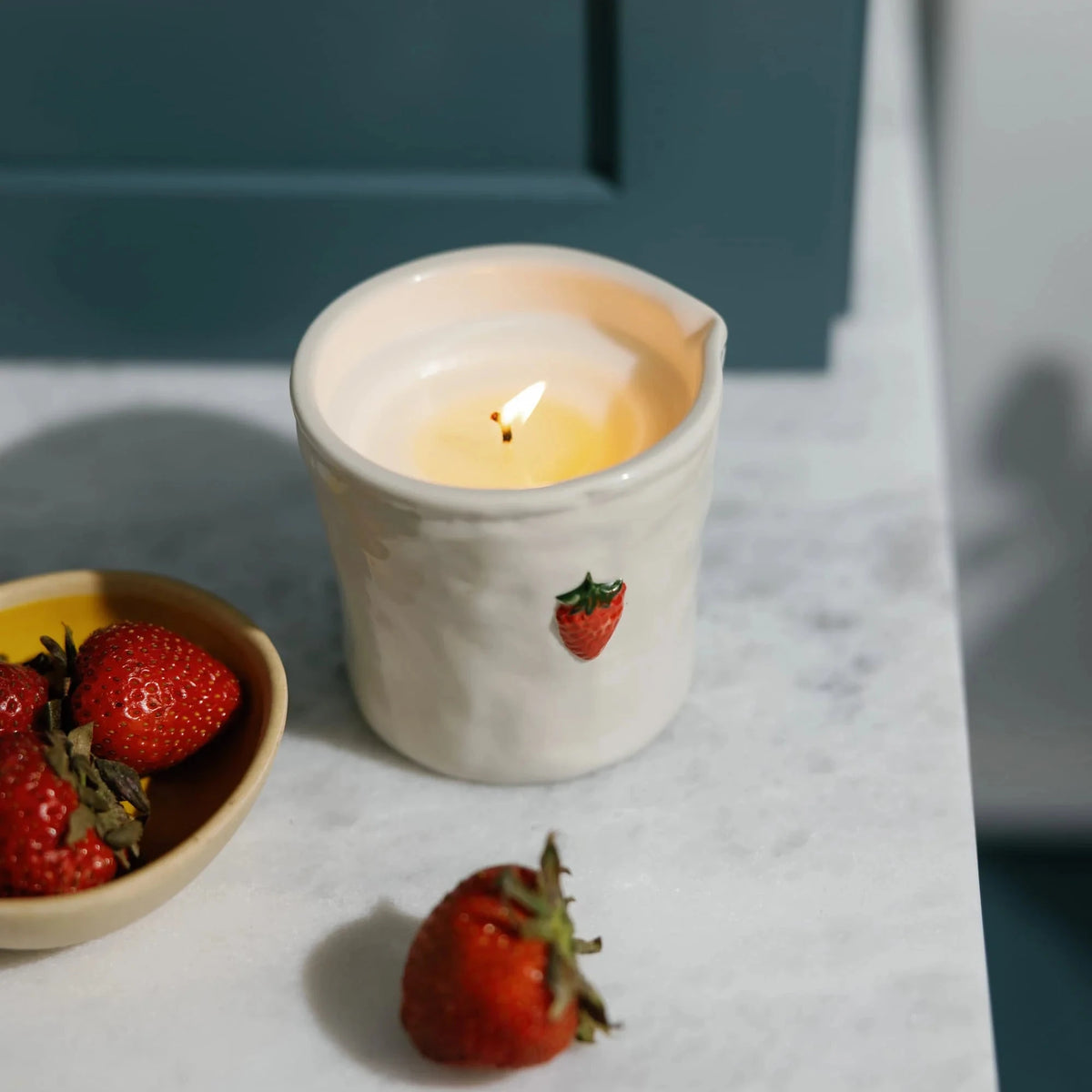 Candle in a white container with strawberry design on a marble surface next to strawberries.