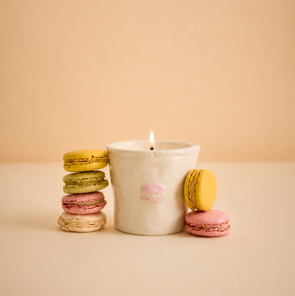 A lit white ceramic candle with a small pink macaron embossed on the front, styled with colorful macaron cookies on a warm beige background.