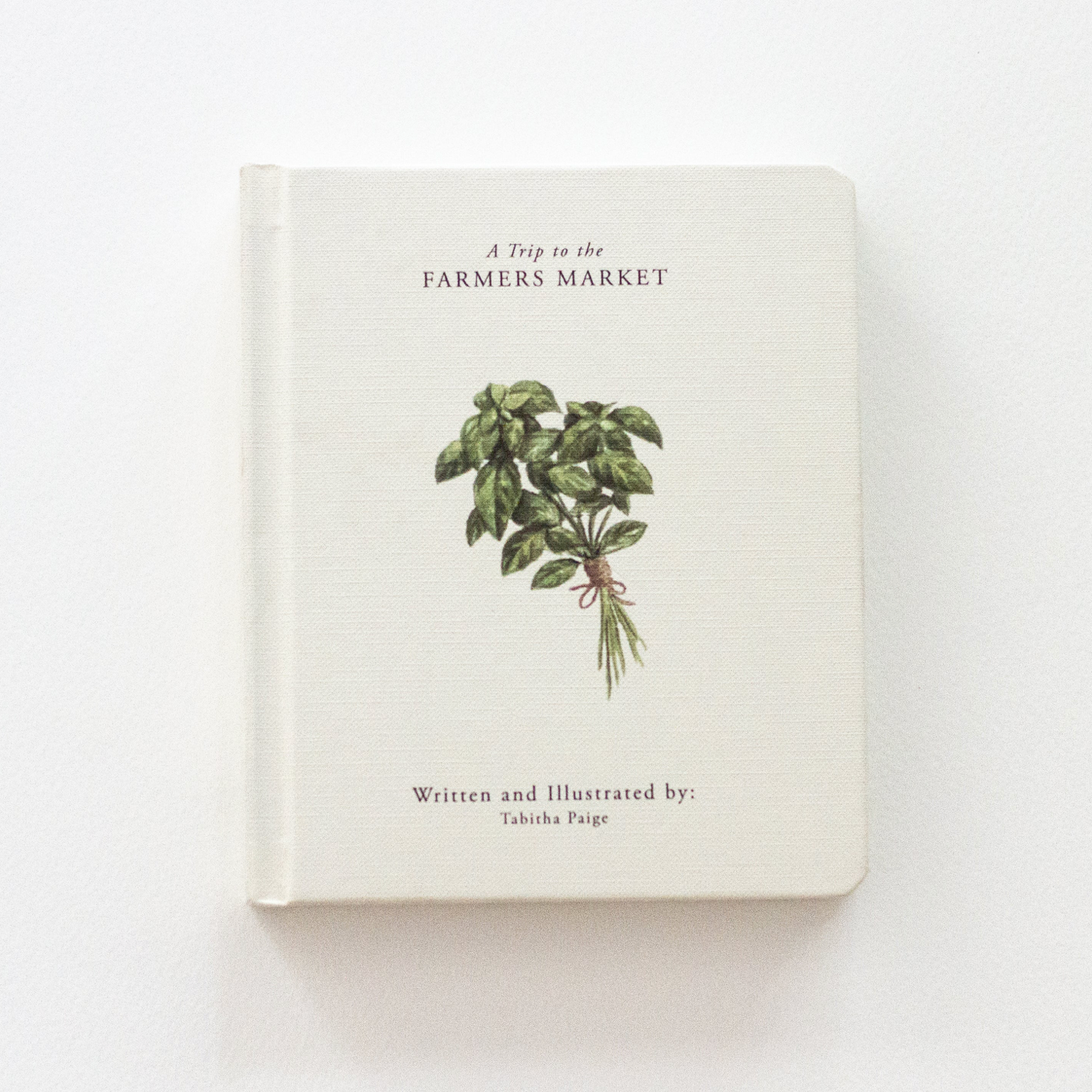 A hardcover book titled 'A Trip to the Farmers Market' by Tahicha Paige, featuring a cream-colored textured cover with an illustration of a green herb sprig in the center.