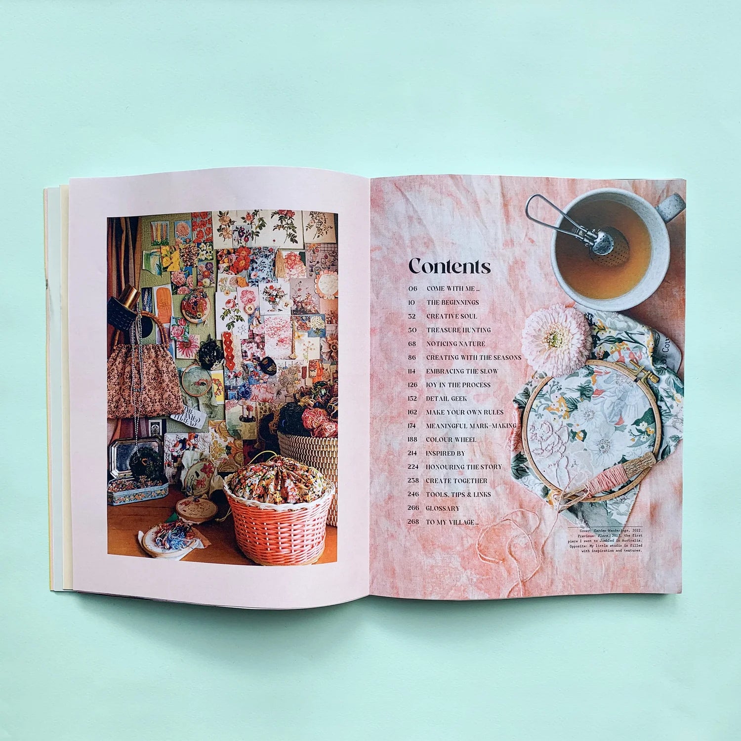 An open book laid flat on a mint green background, showing a colorful interior photograph on the left page and a pink table-of-contents page on the right with a teacup and floral plate.